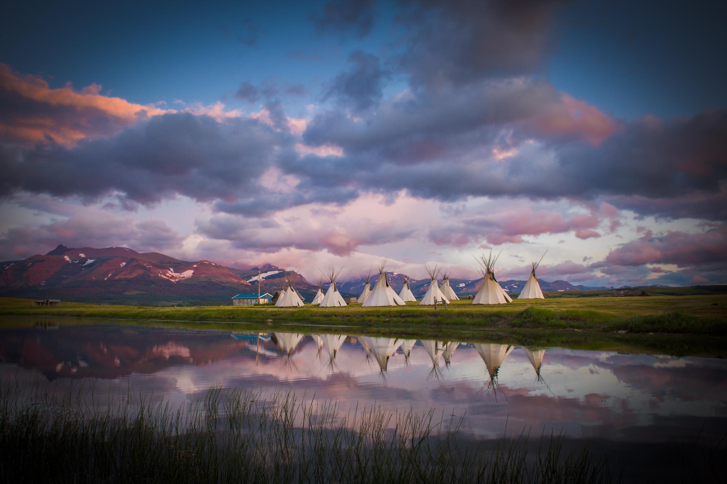 A landscape scene featuring a mountain range with snow patches in the background, a serene lake reflecting the sky, grass in the foreground, and several white teepees arranged in a row, set against a cloudy sky at sunset or sunrise.