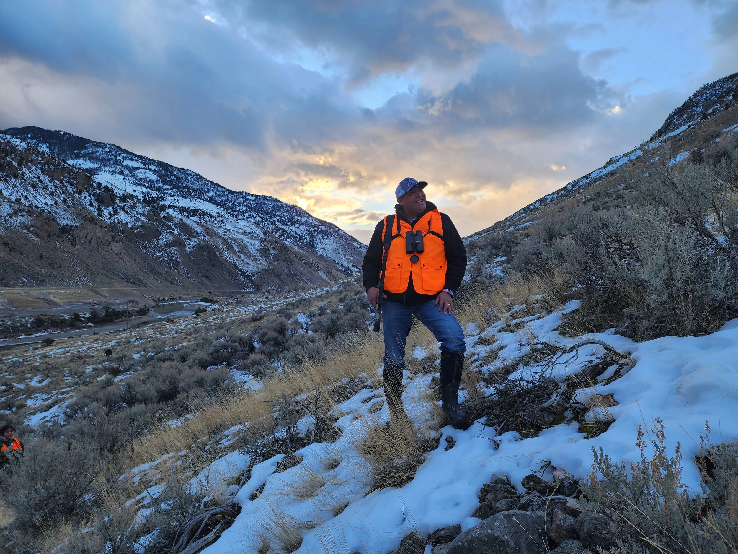 A man standing on a snowy hillside during sunset, wearing an orange vest, black jacket, gray cap, and binoculars around his neck, with mountains and a cloudy sky in the background.