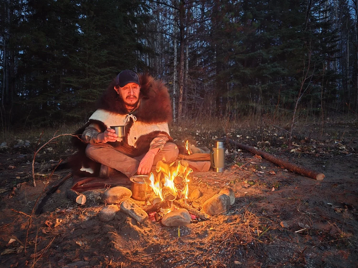A person sitting by a campfire in a forest at dusk, holding a cup, with a thermos nearby, wearing warm clothing with a fur-lined cape, surrounded by trees and natural outdoor elements.