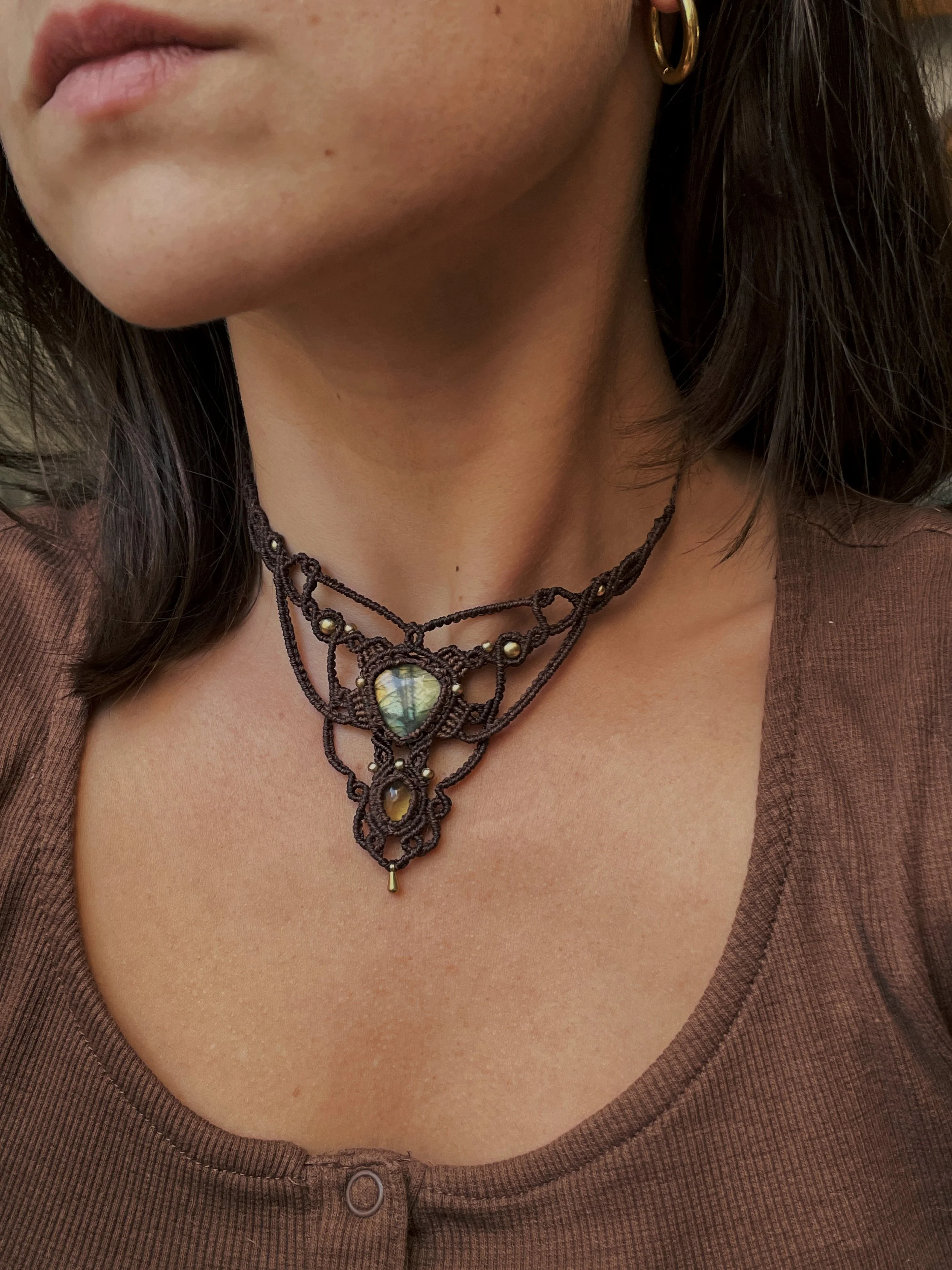 Close-up of a woman's neck and chest area, wearing a brown button-up top with the top button undone, gold hoop earrings, and a layered black necklace with a large iridescent gemstone centerpiece.