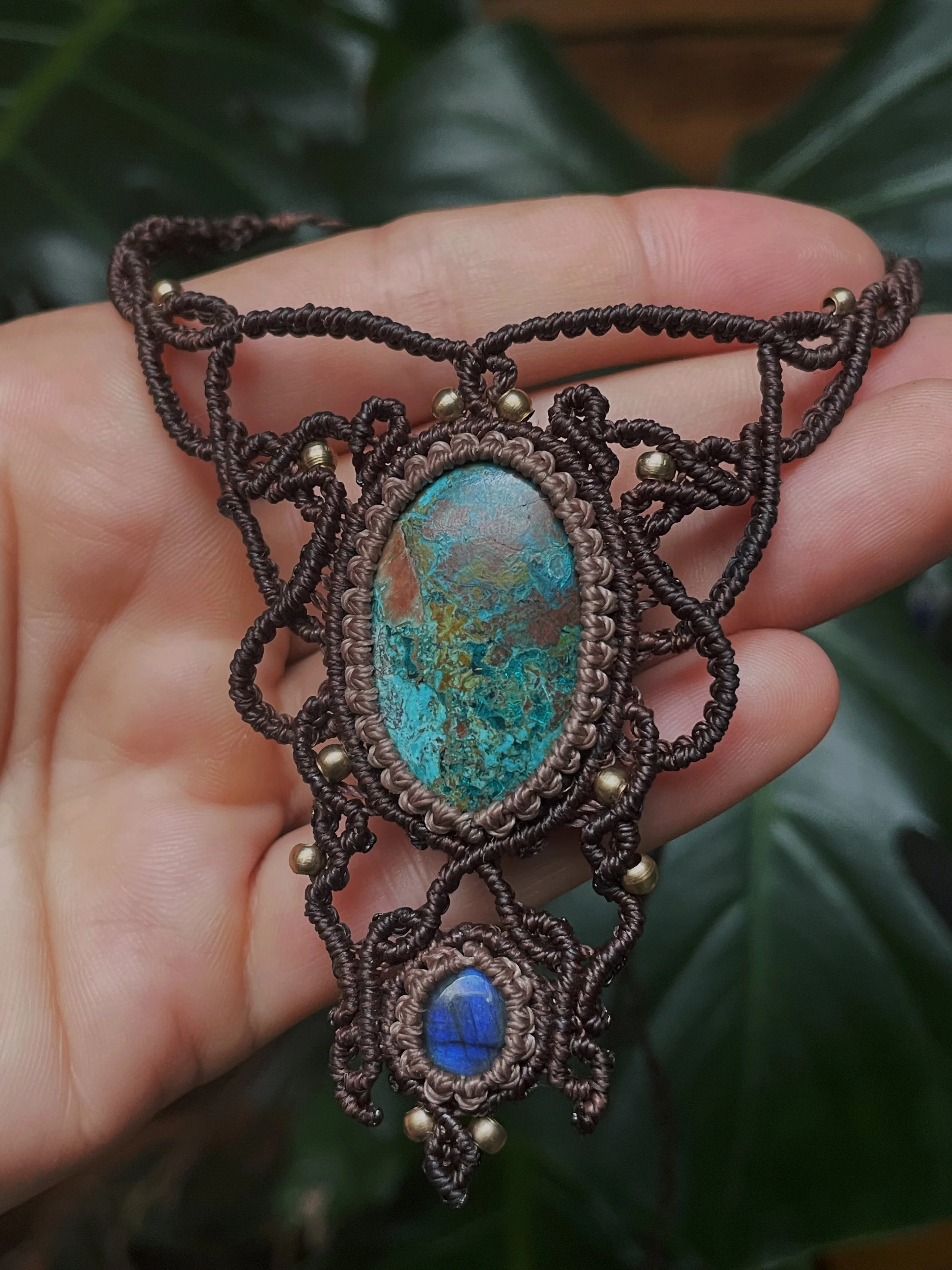 Hand holding a woven necklace with large oval turquoise stone and small blue bead, surrounded by intricate brown macrame and gold beads, with green foliage in the background.