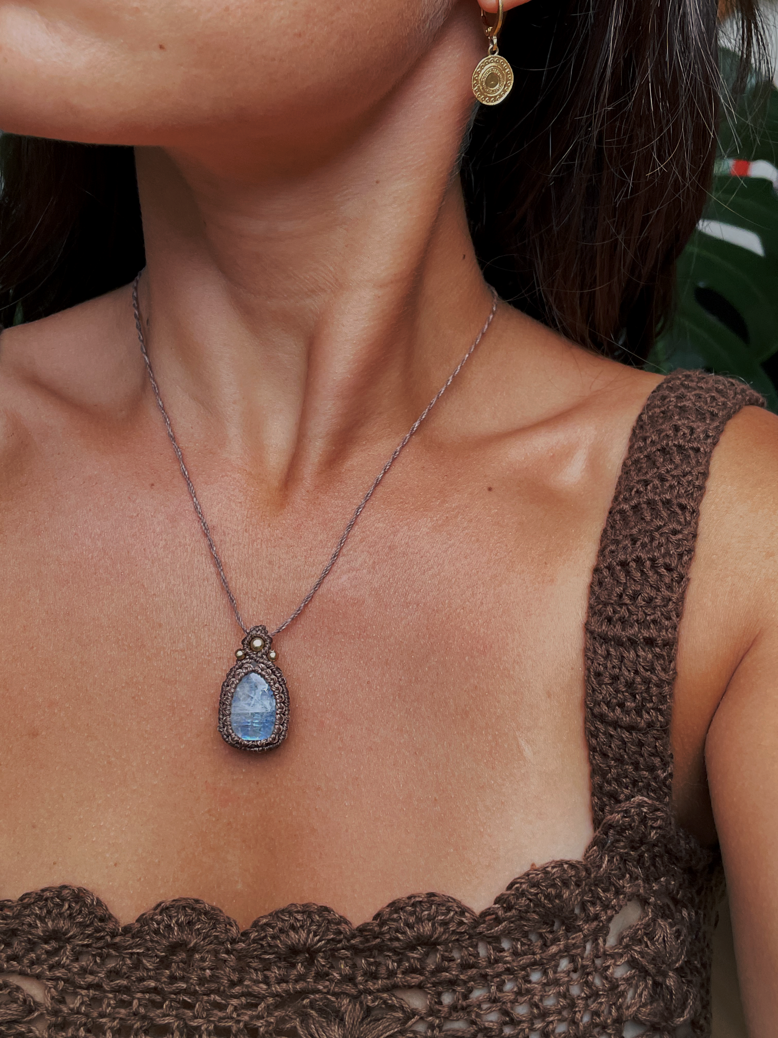 Close-up of a woman wearing a moon stone necklace and gold earrings, with a knitted brown top and a background of green leaves.