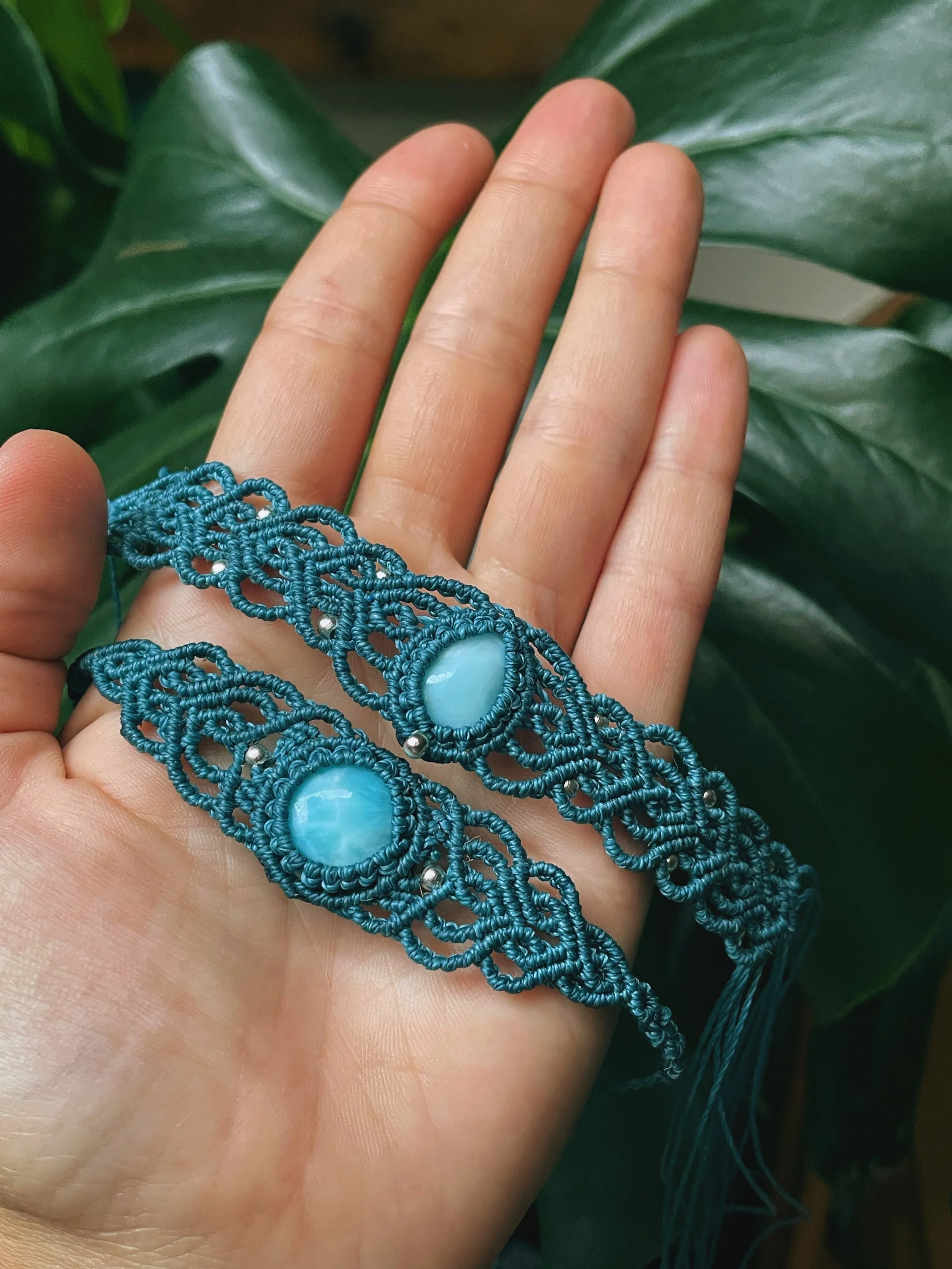 Close-up of a hand wearing a blue lace bracelet with large blue stones, against green leaves in the background.