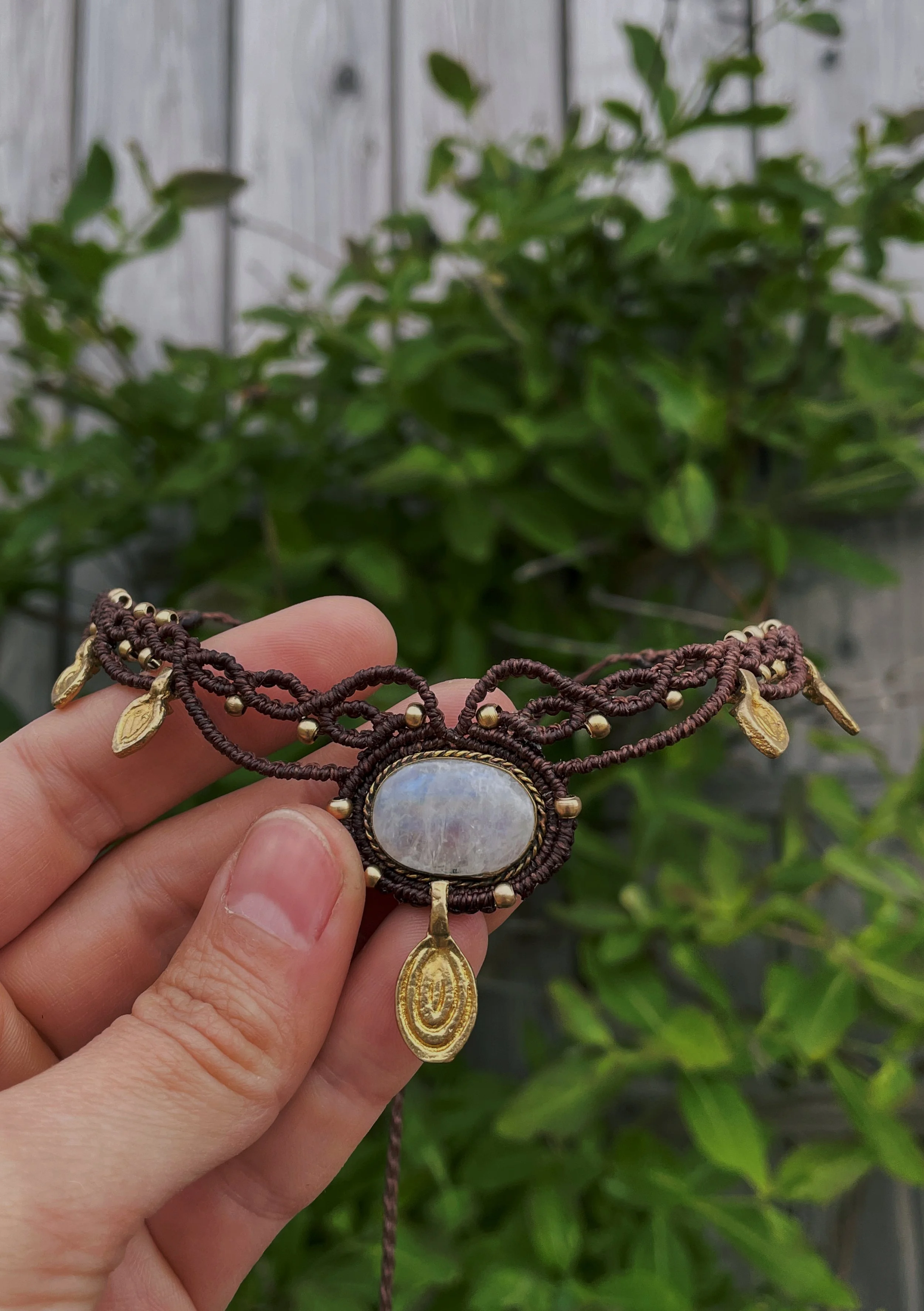 Person holding a necklace with a large oval gemstone, gold charms, and a gold oval pendant with intricate designs, against a background of green foliage and a gray wooden fence.