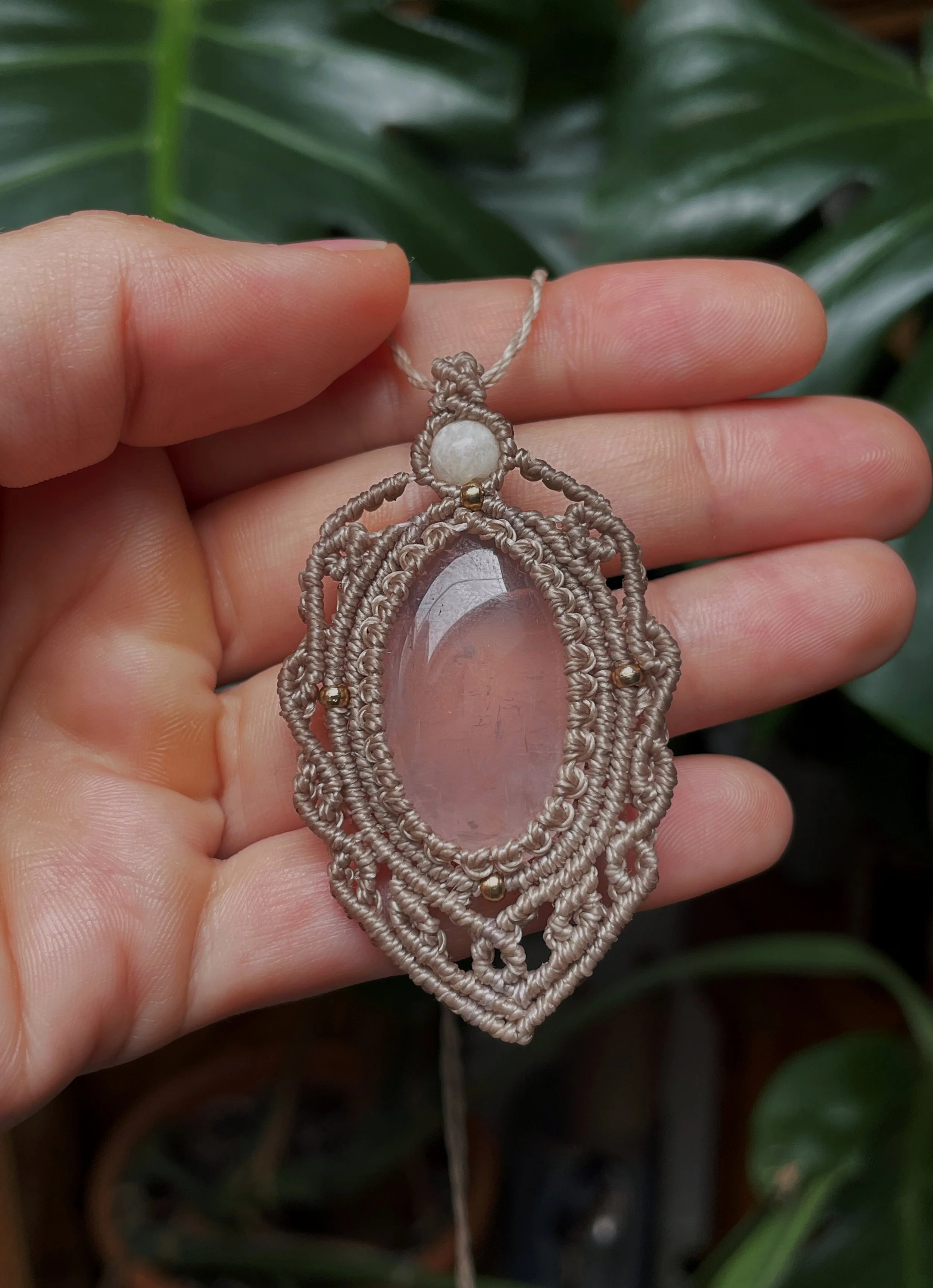 Hand holding a decorative necklace with a large pink stone and intricate beige woven design, with green foliage in the background.
