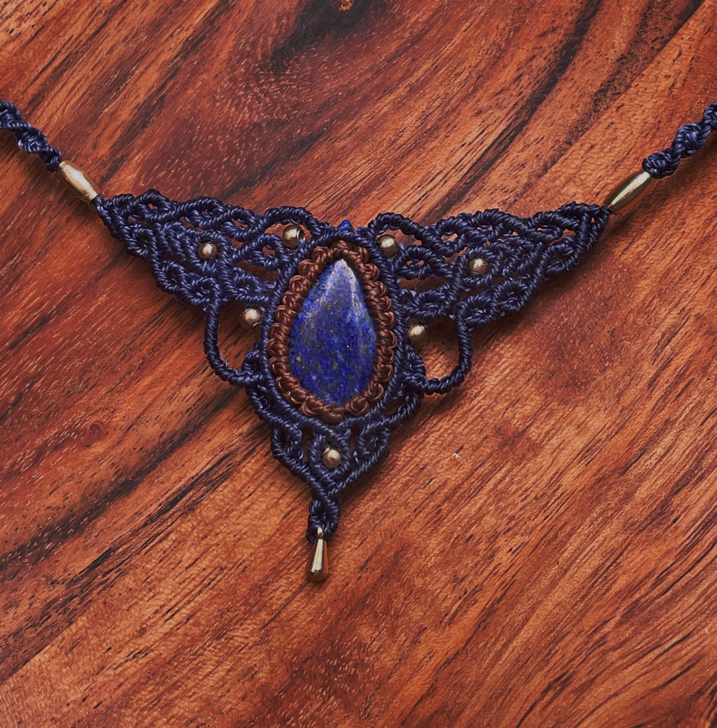 Close-up of a handcrafted macrame necklace with a large lapis lazuli stone pendant, gold beads, on a wooden surface.