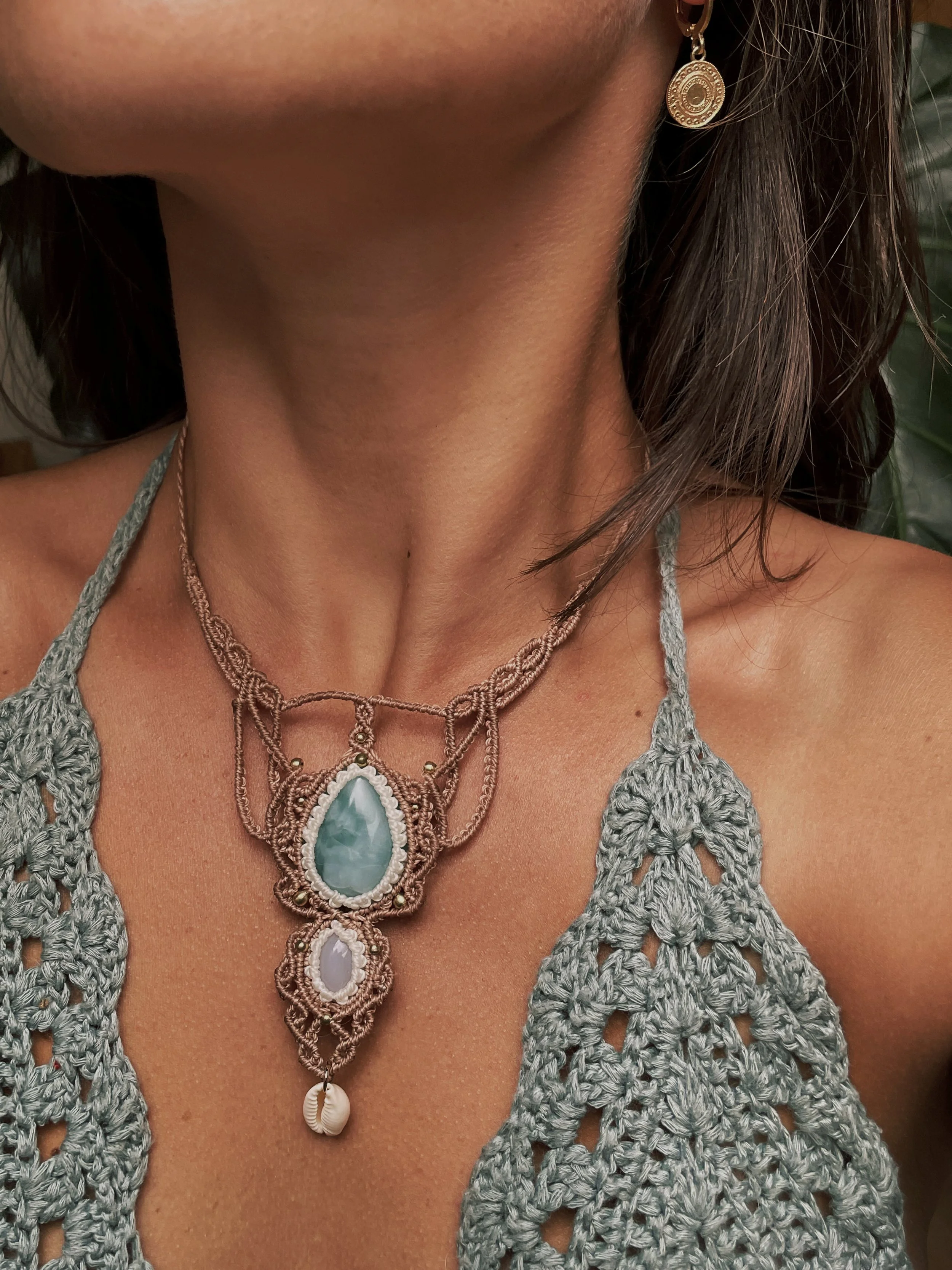 Close-up of a woman's neck wearing a layered necklace with large gemstones, a shell charm, earrings, and a gray crocheted top.