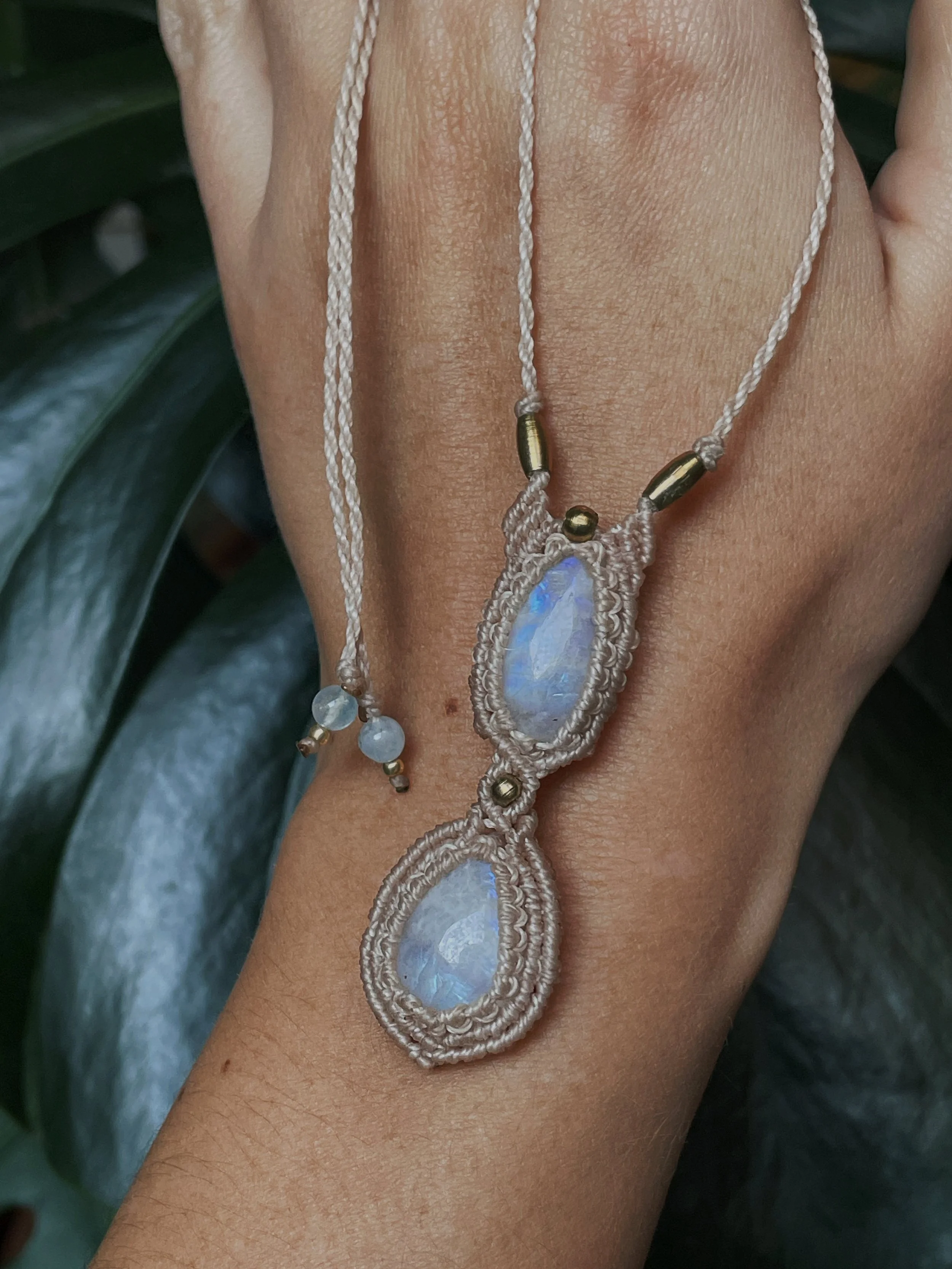 Close-up shot of a person's arm adorned with a macrame necklace with an oval-shaped opal pendant and small beads, set against dark green foliage.