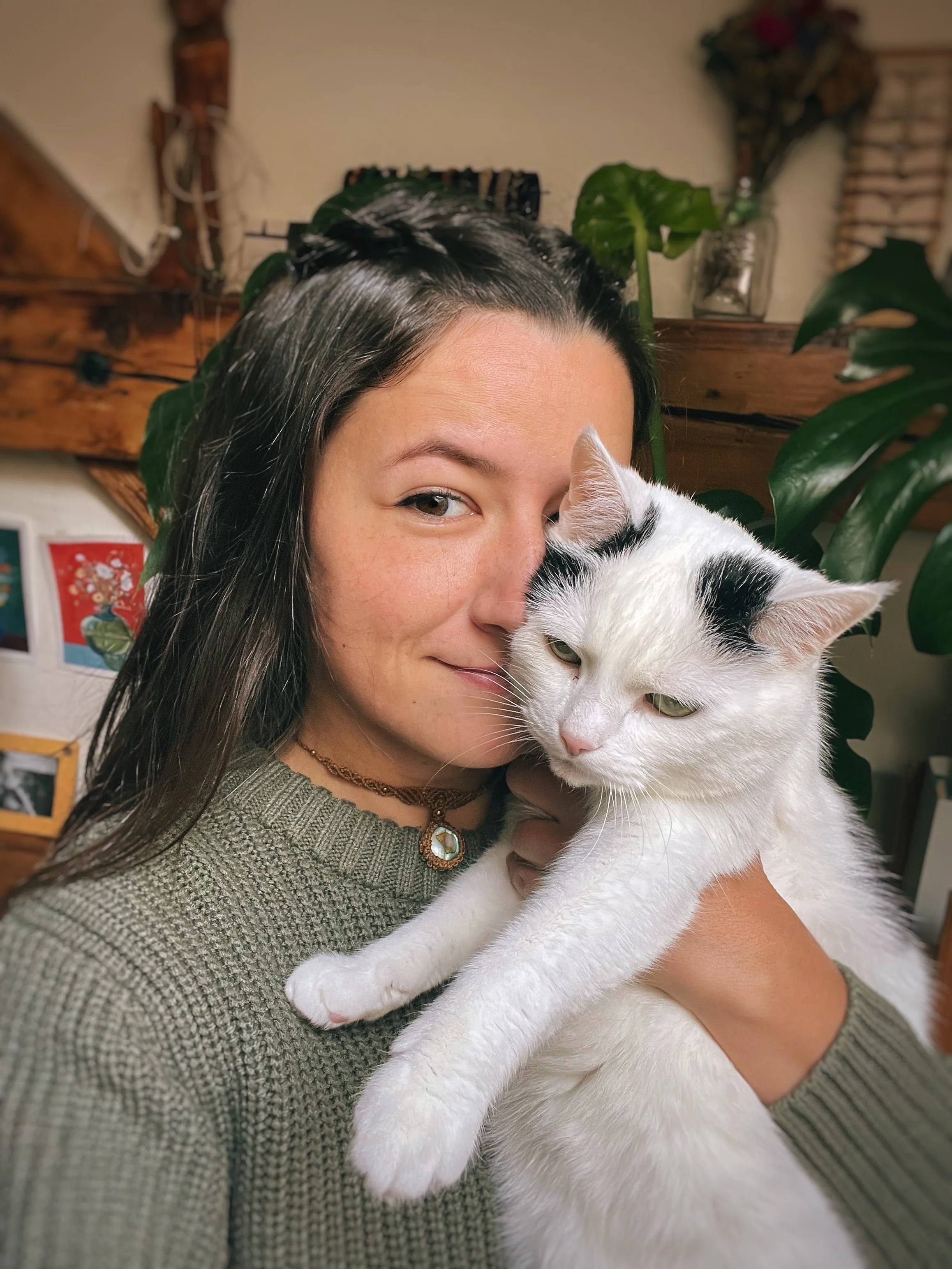 A woman with long dark hair and a grey knitted sweater is holding a white cat with black markings close to her face. The woman is smiling slightly and has a necklace with a pendant. The background includes a wooden shelf with green plants, a jar, and some wall decorations.
