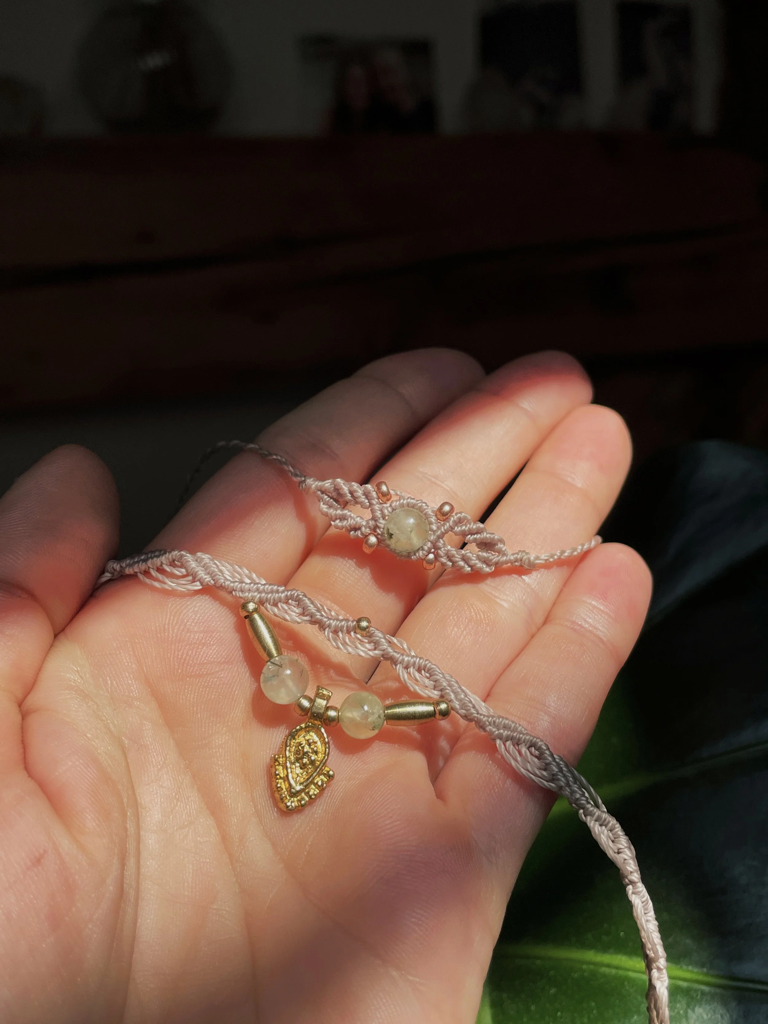 A hand holding a pink or beige macrame bracelet with pearl beads and a gold religious pendant. The background is dark and blurry.