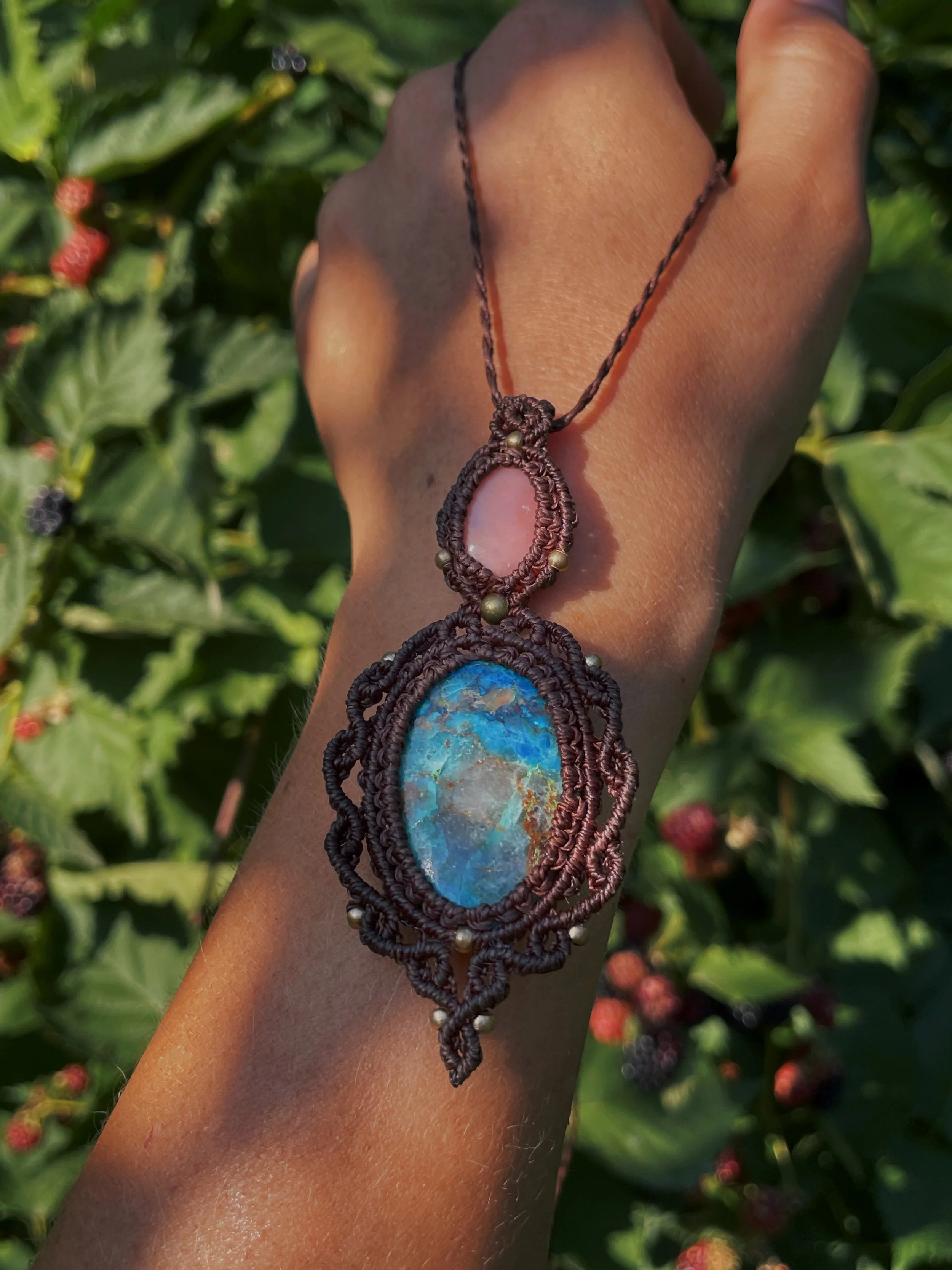 A person's hand with a macrame necklace featuring a large, oval, iridescent stone, held up against a background of green foliage and blackberries.