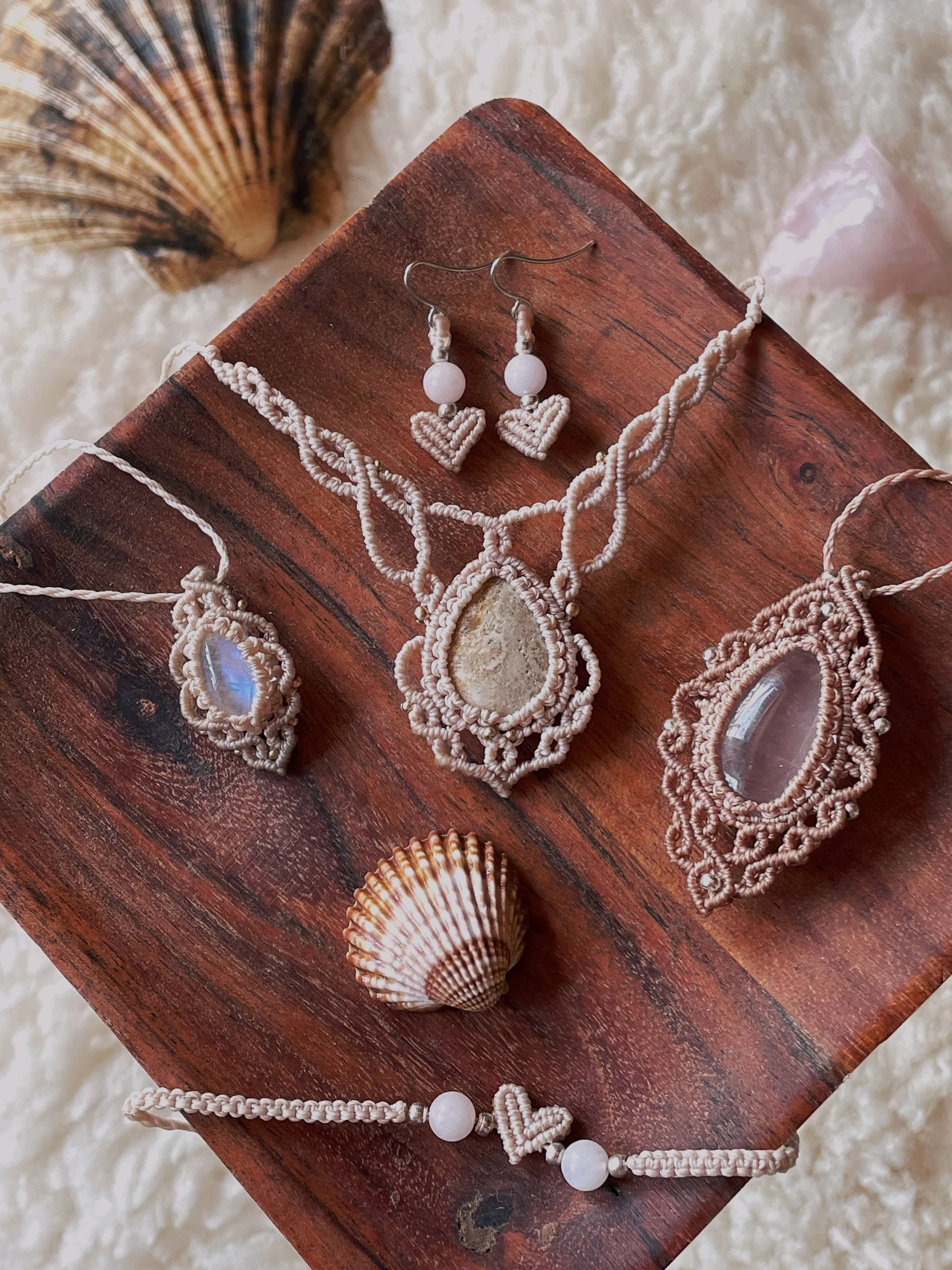 Beach-themed jewelry set including a necklace, earrings, and a bracelet with macrame designs and seashell pendants, displayed on a wooden surface with shells in the background.