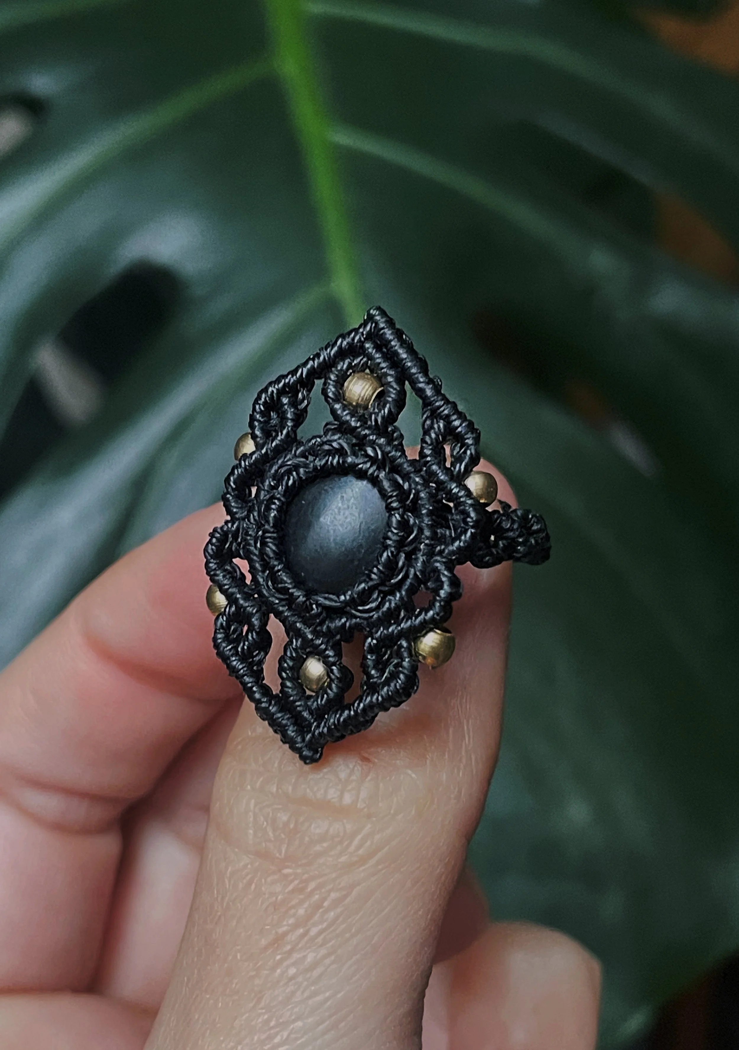 Close-up of a person holding a black macrame ring with gold beads, with green foliage in the background.