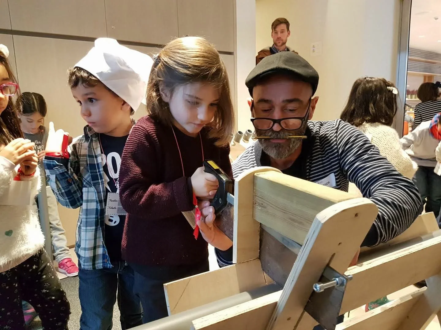 Hombre con barba, gafas, y boina, enseñando a niños en actividad con una tabla de madera y herramientas en ambienten interior.