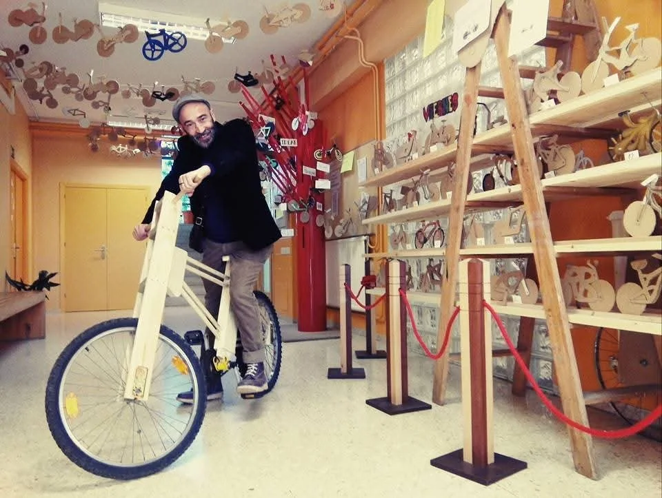 Hombre en una rueda de bicicleta de madera dentro de una sala decorada con obras de arte en madera y objetos decorativos en el techo.