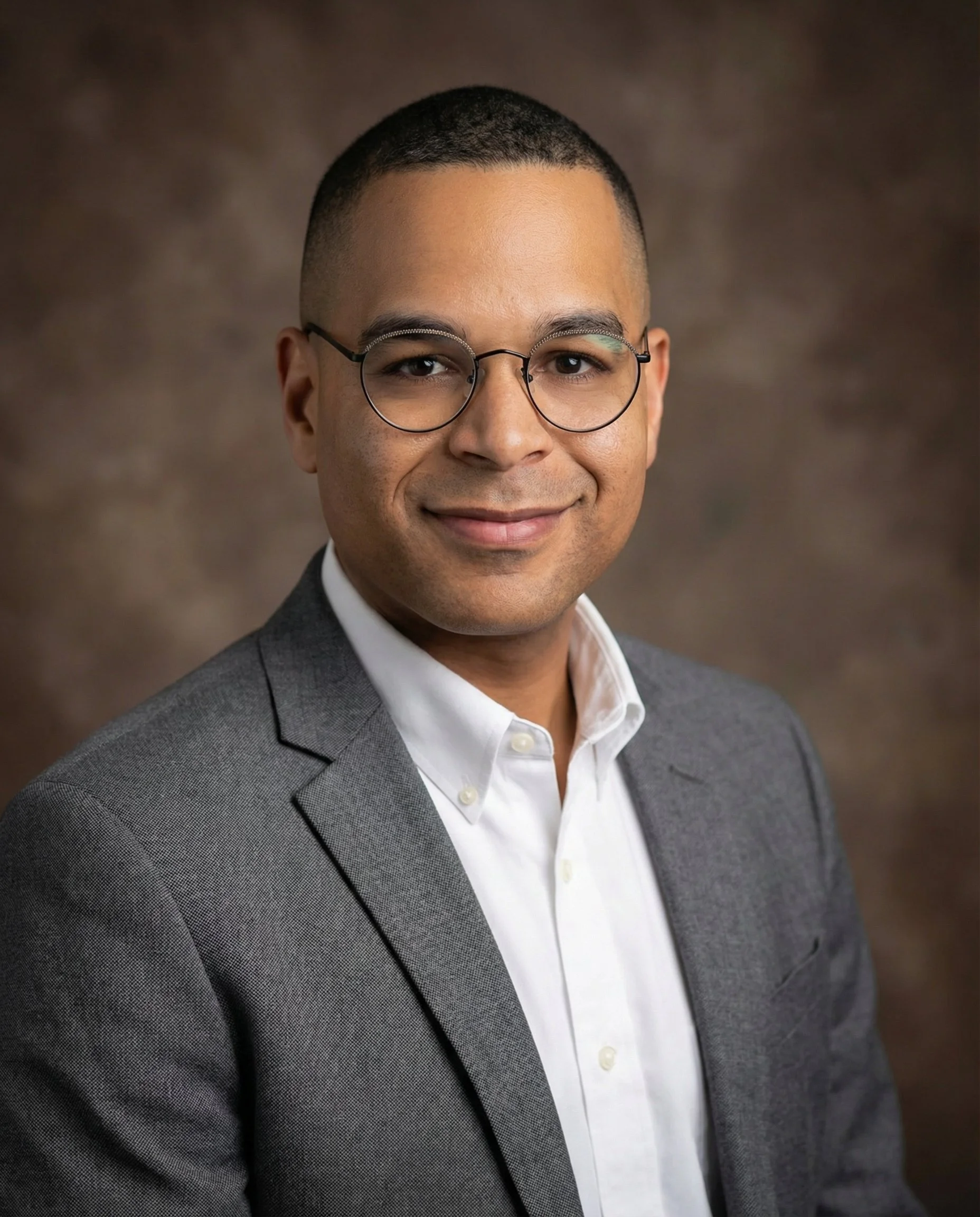 Headshot of a Simeon Ngweso, Berwick Urologist,  with short hair and glasses, wearing a gray suit jacket and white dress shirt, smiling against a brown background.