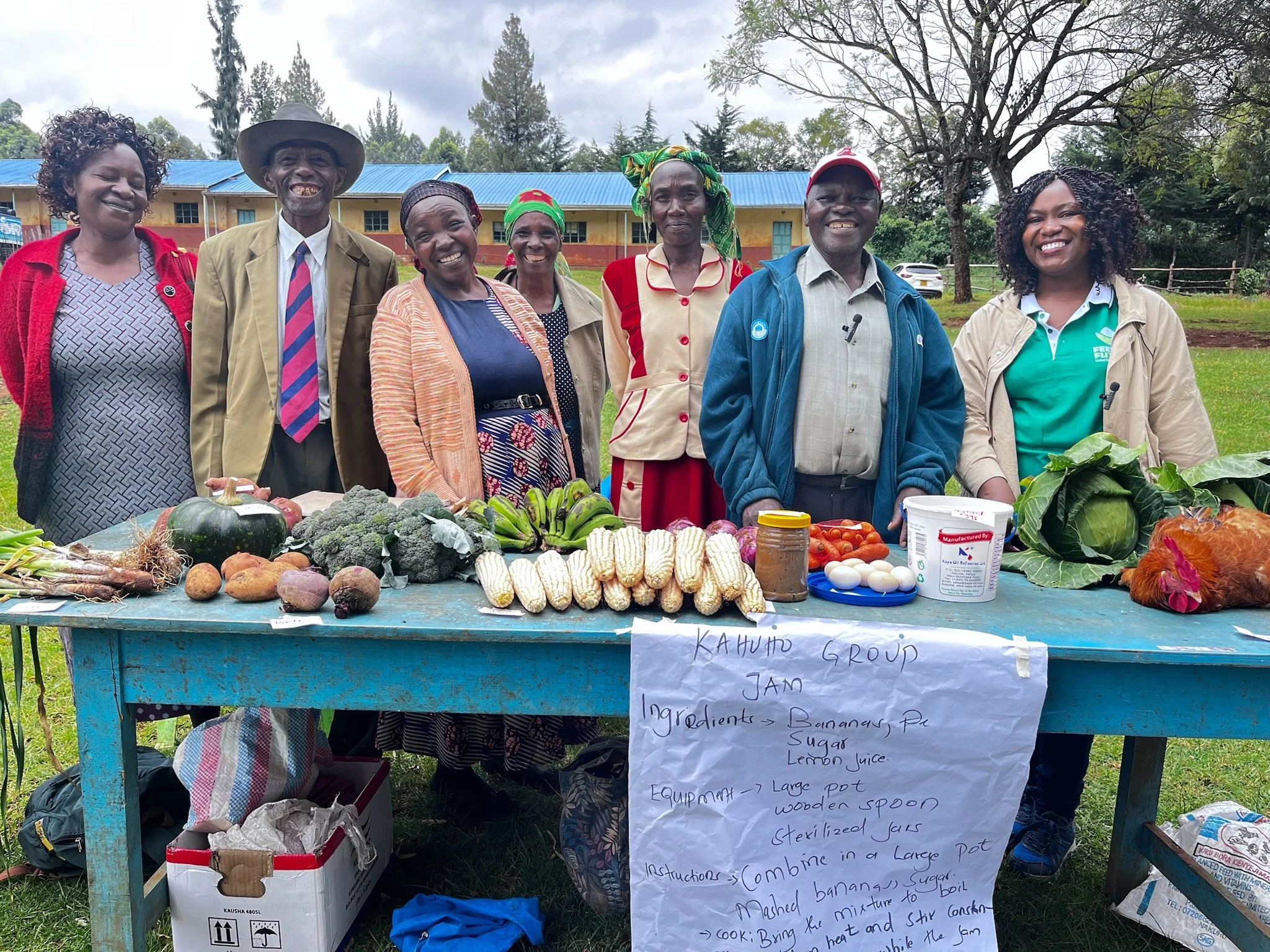 A group of seven people standing behind a table with vegetables and fruits, outdoors in a green area with trees and a building in the background. They are smiling and appear to be at a market or community event.