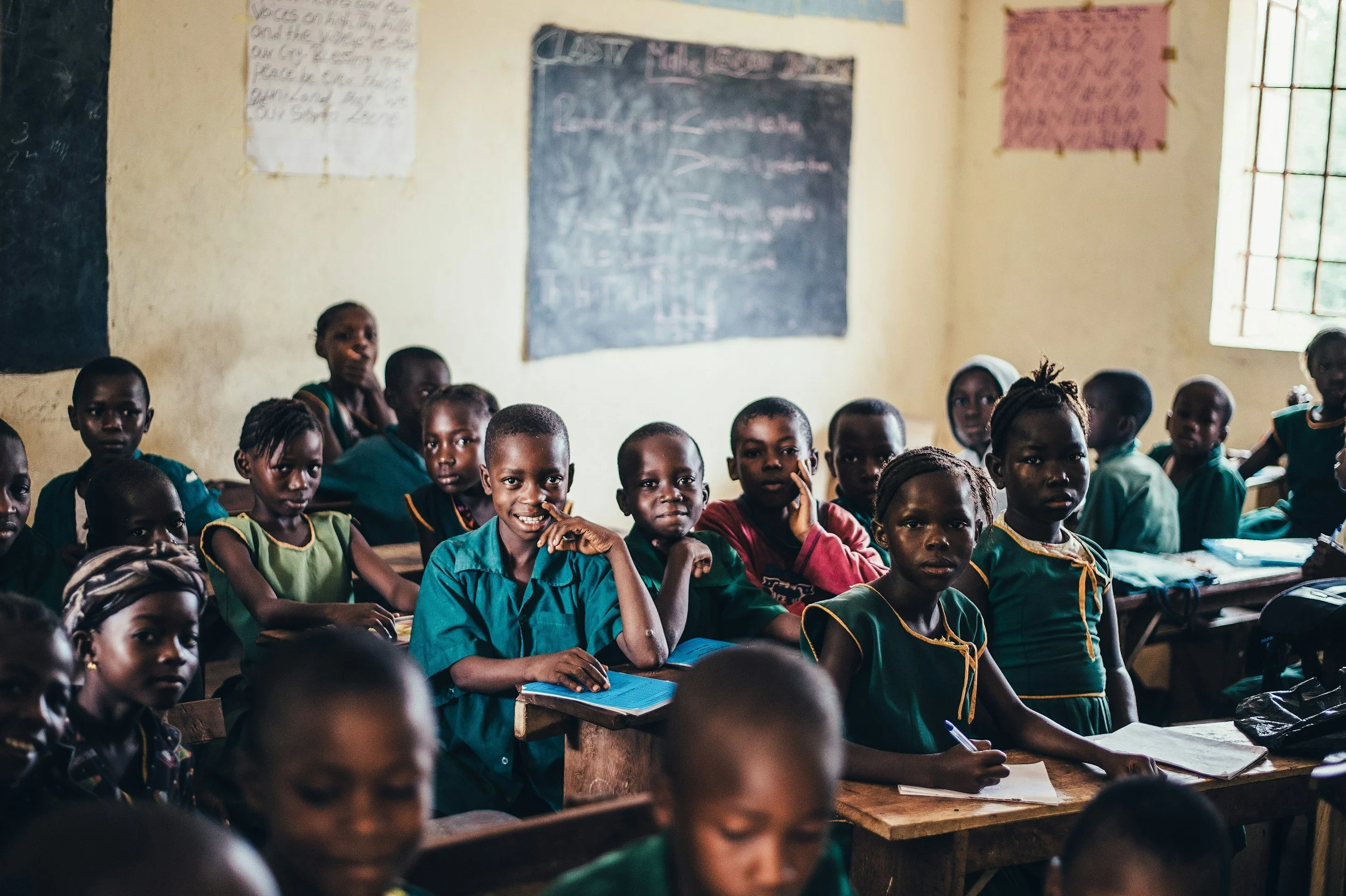 A classroom filled with young students sitting at desks, looking towards the camera. There are blackboards and posters on the wall behind them, with natural light coming through a window on the right.