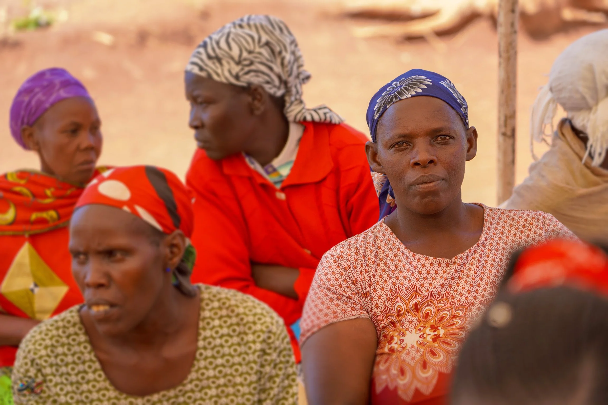 Group of women wearing colorful headscarves and patterned dresses, sitting outdoors in a rural setting.