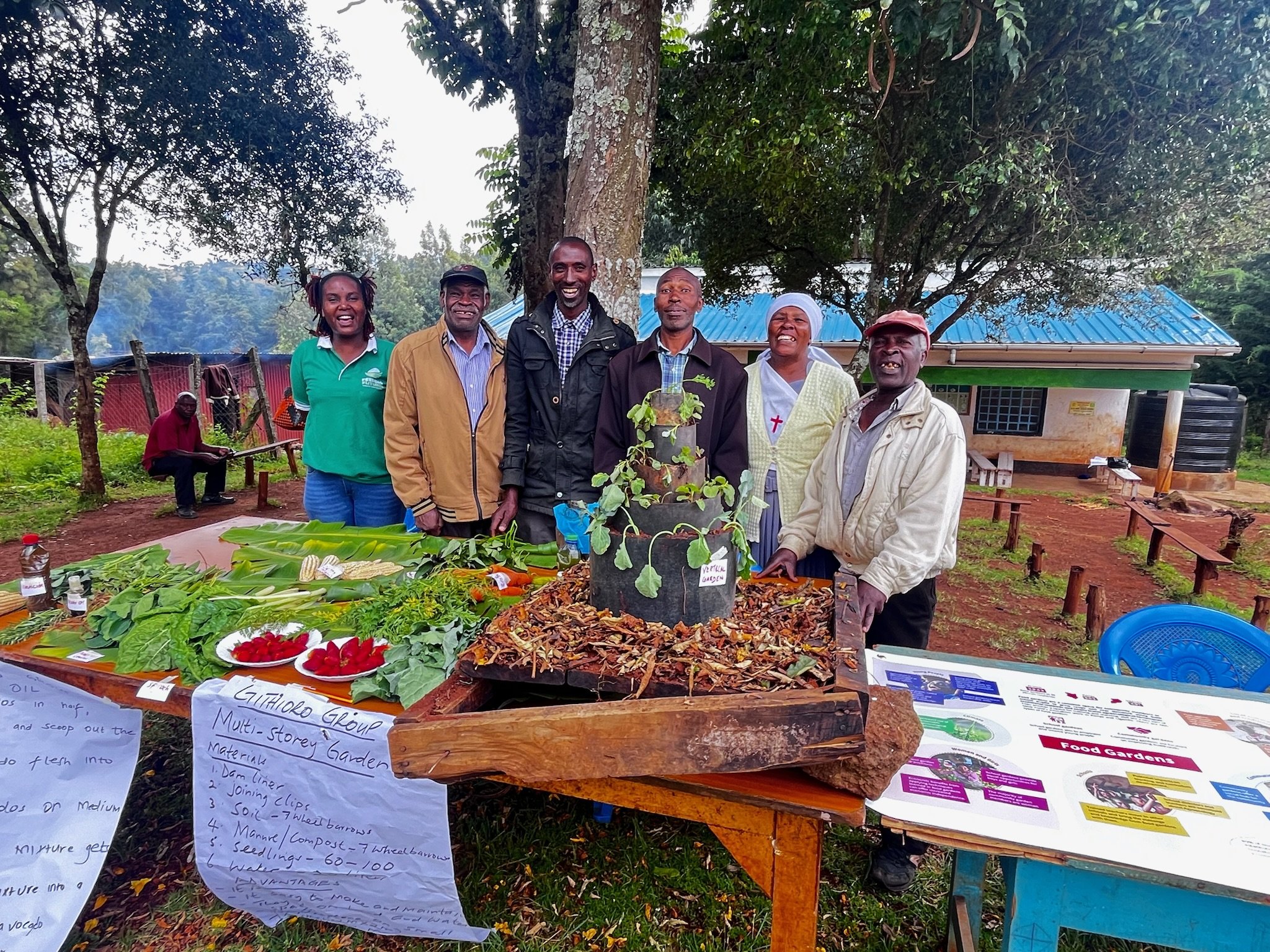 Group of six people standing outdoors around a table with various plants and informational posters. They are smiling and appear to be at a community or educational garden event. There are trees and a small building in the background.