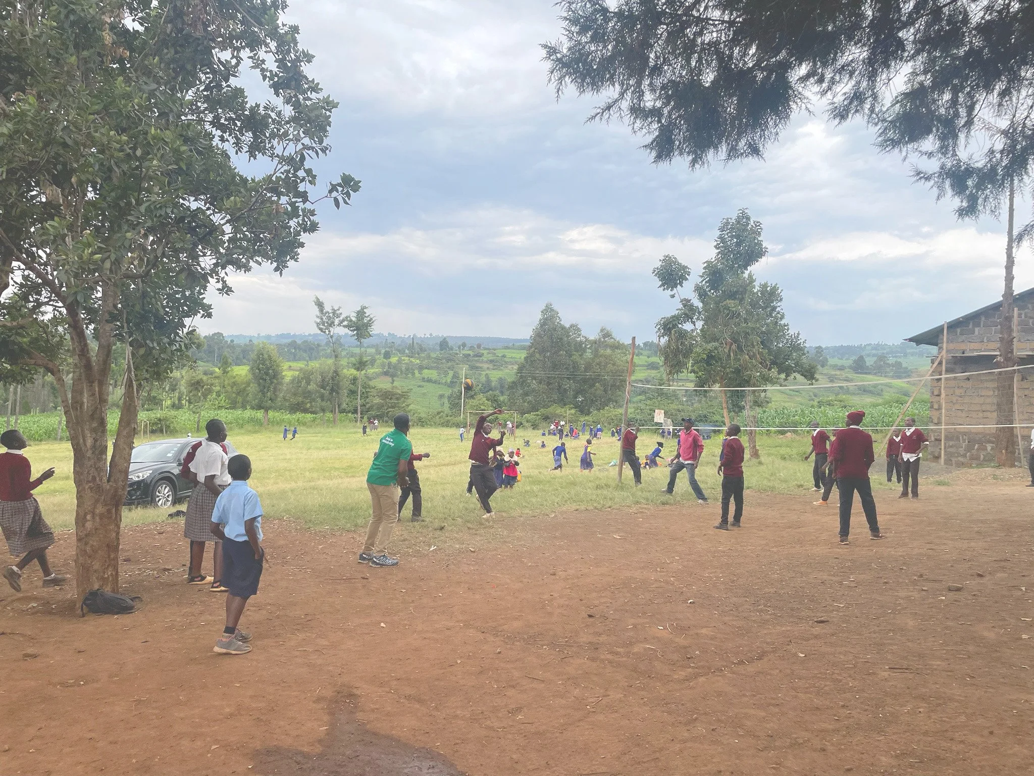 Children and adults playing soccer on a grassy field in a rural area with trees and hills in the background under a cloudy sky.