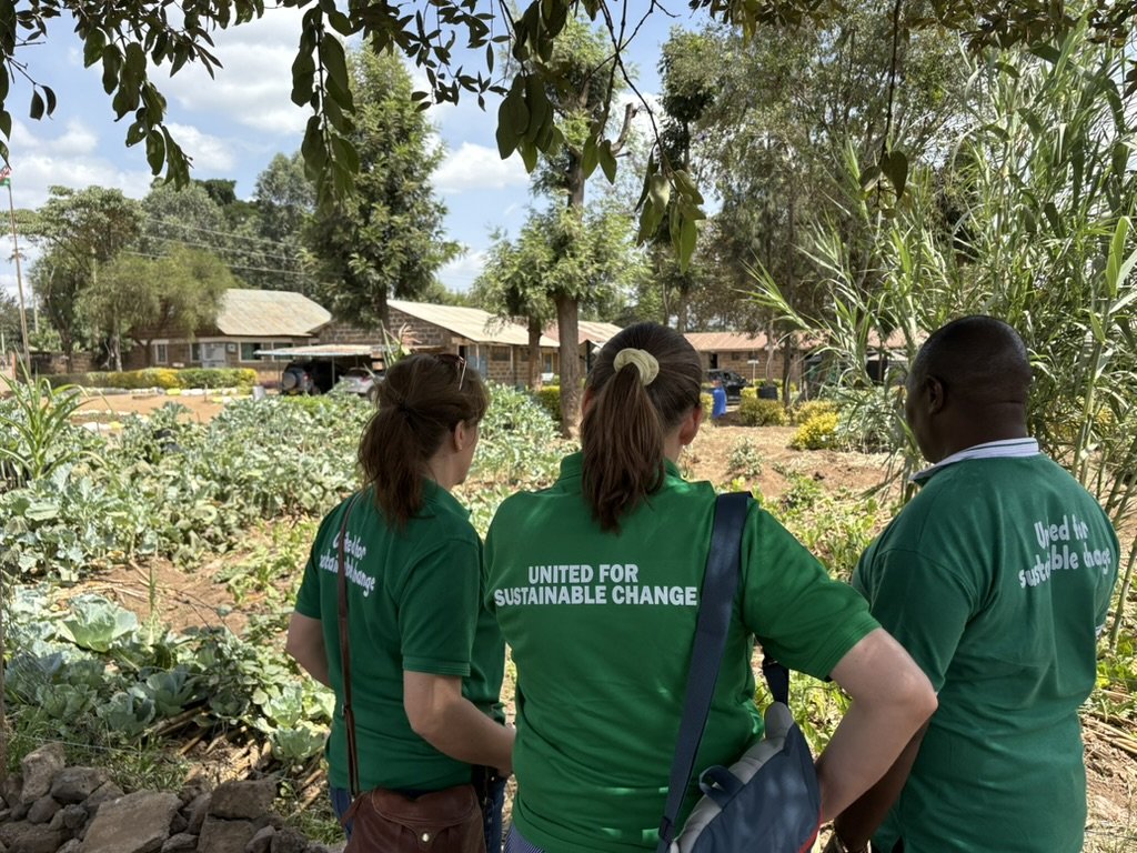 Three people in green shirts with the message "United for Sustainable Change" on the back, standing outdoors in a garden with various plants and trees, engaging in a conversation.