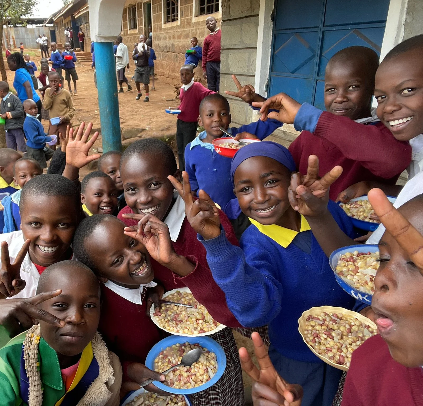 Group of children smiling and holding bowls of food outdoors, some children making peace signs, in front of a building with other children and adults in the background.
