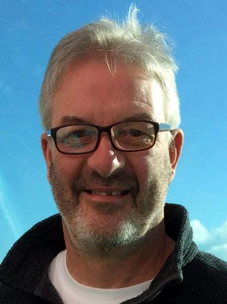 A middle-aged man with light-colored hair and a beard, wearing glasses, smiling outdoors with a blue sky and some clouds in the background.