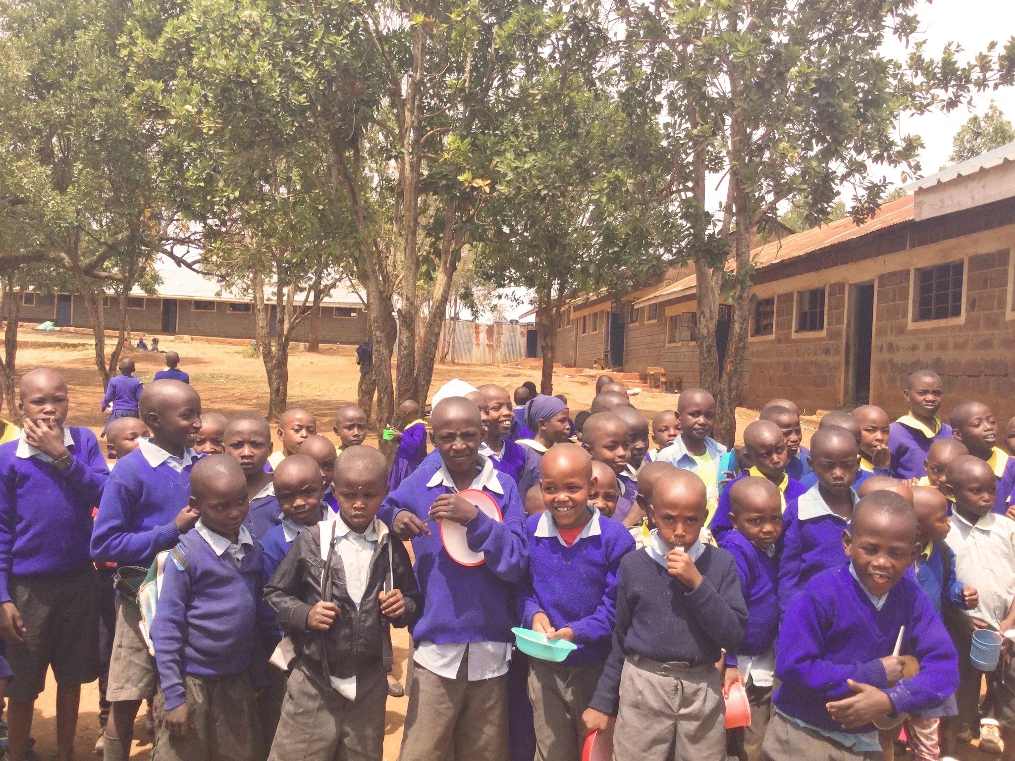 Group of children and a few adults standing outdoors near trees and a brick building, some children holding containers and looking towards the camera.