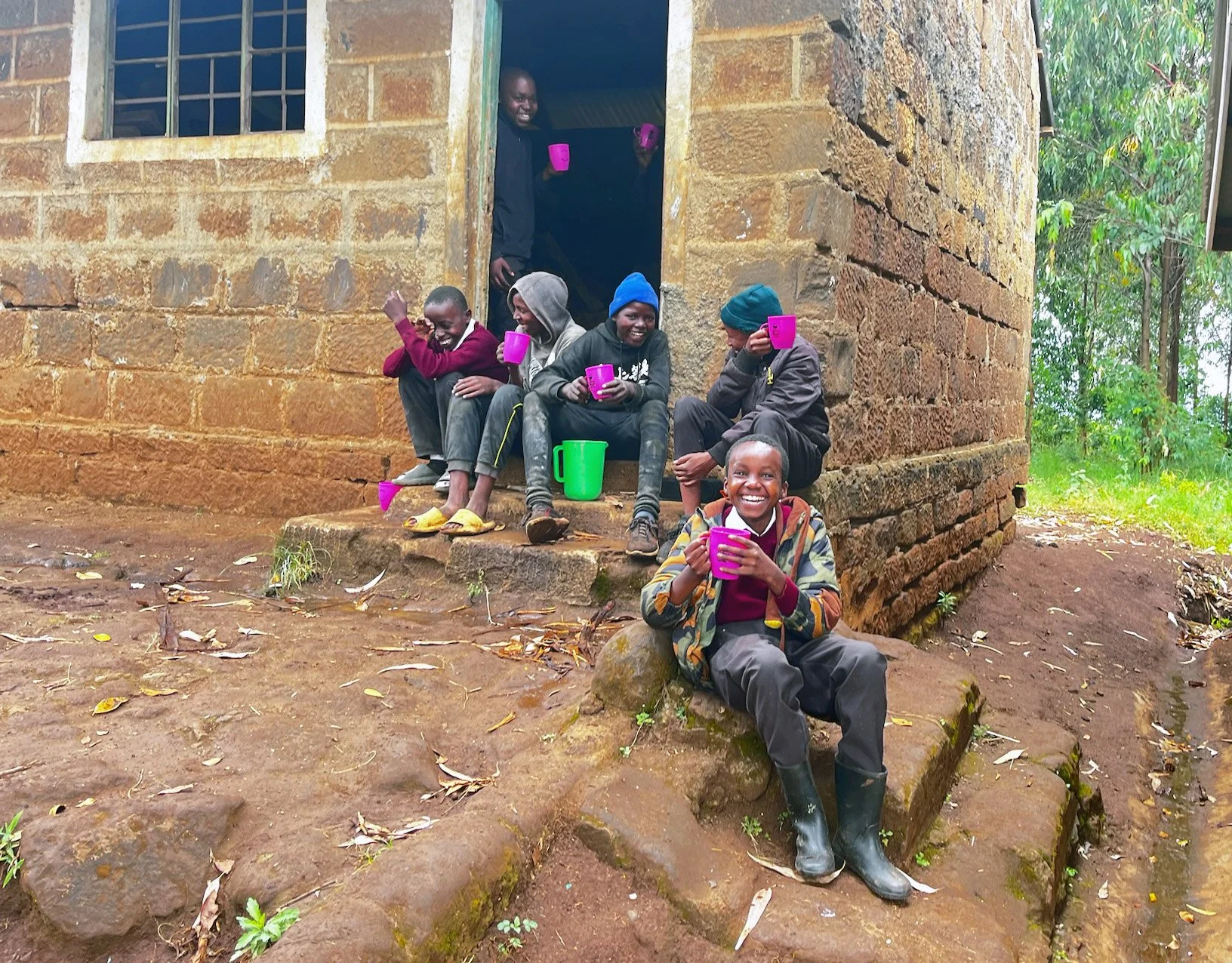 Youth sitting on stone steps outside a brick building, holding colorful cups, with one boy in front sitting on a rock, smiling, while another peeks out from the doorway, smiling.