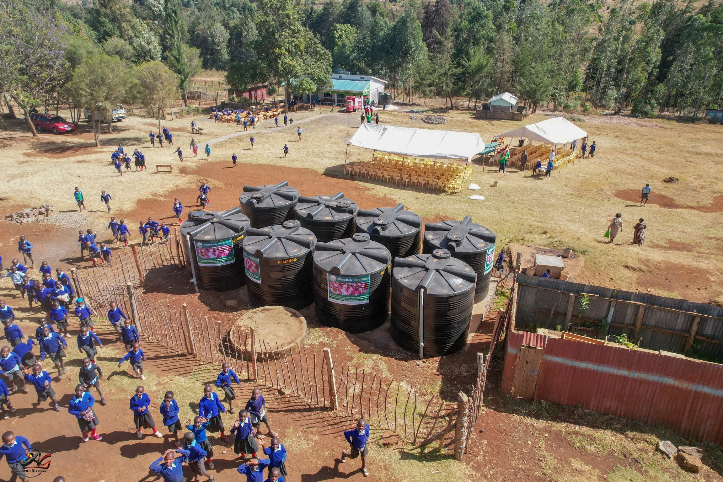 Aerial view of a schoolyard with children in uniform walking and playing on a dirt path, large water tanks, tents with chairs, and buildings surrounded by trees.
