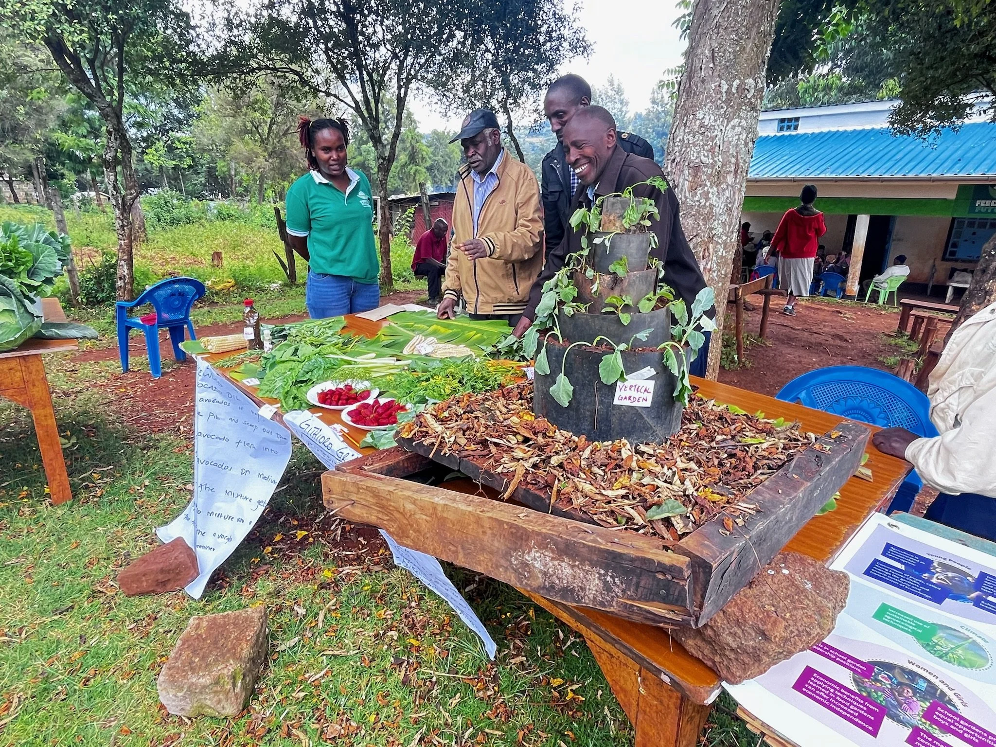 People at a market stall selling vegetables and plants outdoors under trees, with signs and papers on the table.
