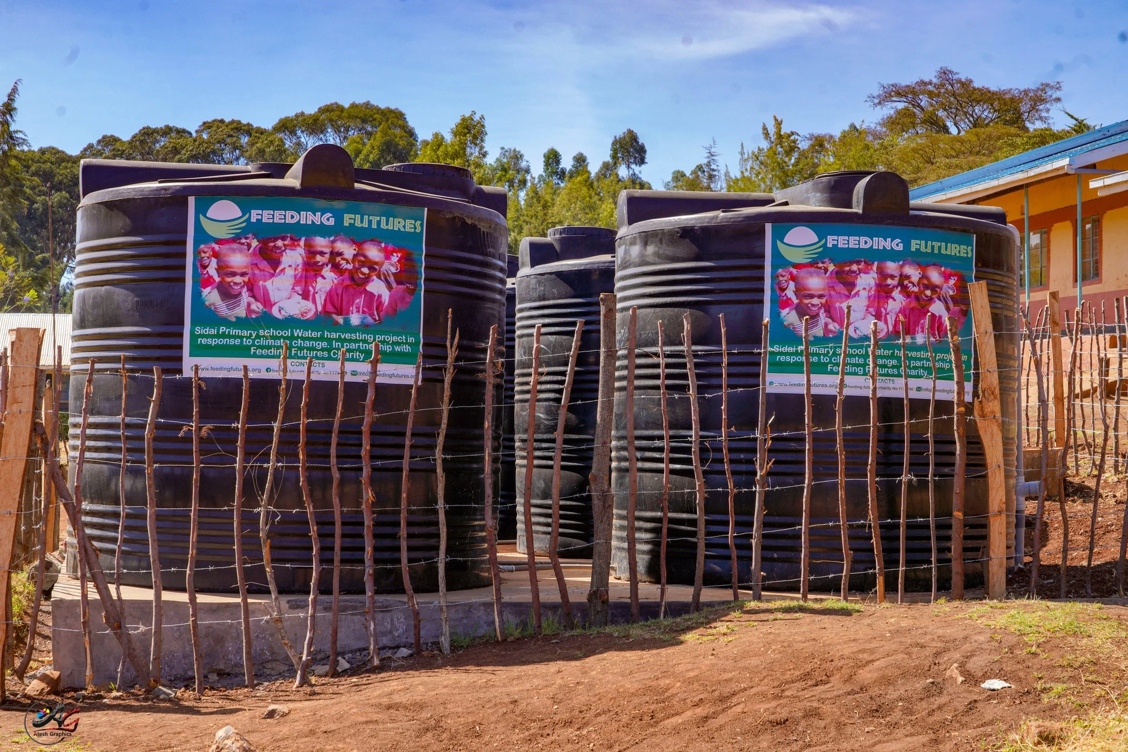 Black water tanks with a poster that reads 'Feeding Futures' and shows children, situated outdoors behind a wooden fence with trees and a building in the background.