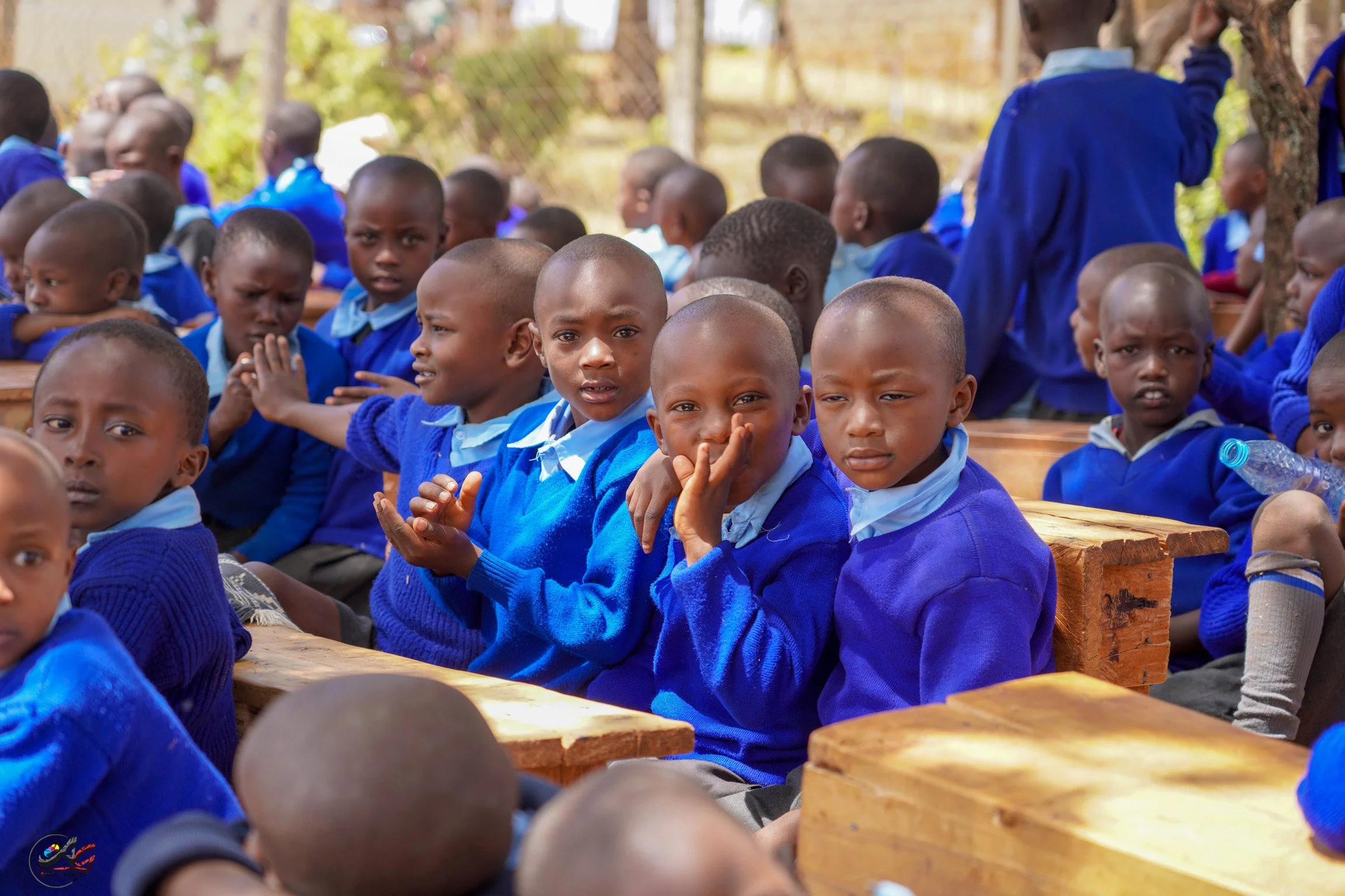 A group of young children sitting outdoors on wooden benches, wearing blue school uniforms, some looking directly at the camera with curious and serious expressions, others looking away or engaging with something outside the frame.
