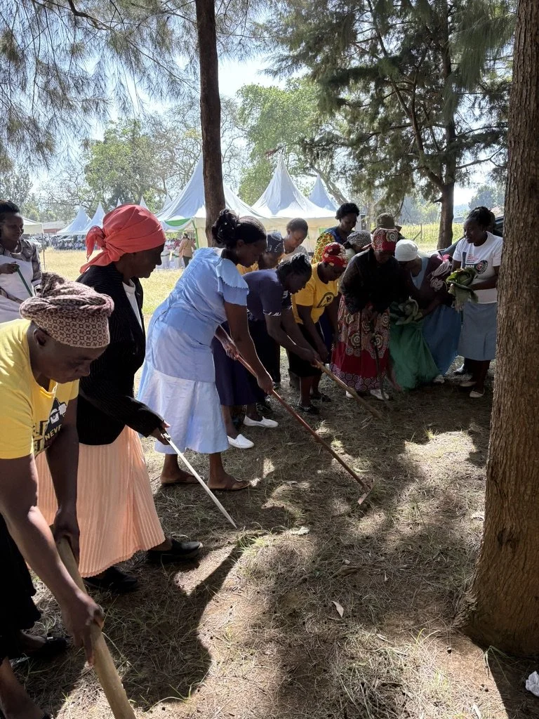 Group of women and girls planting trees in a shaded outdoor area with tents in the background.