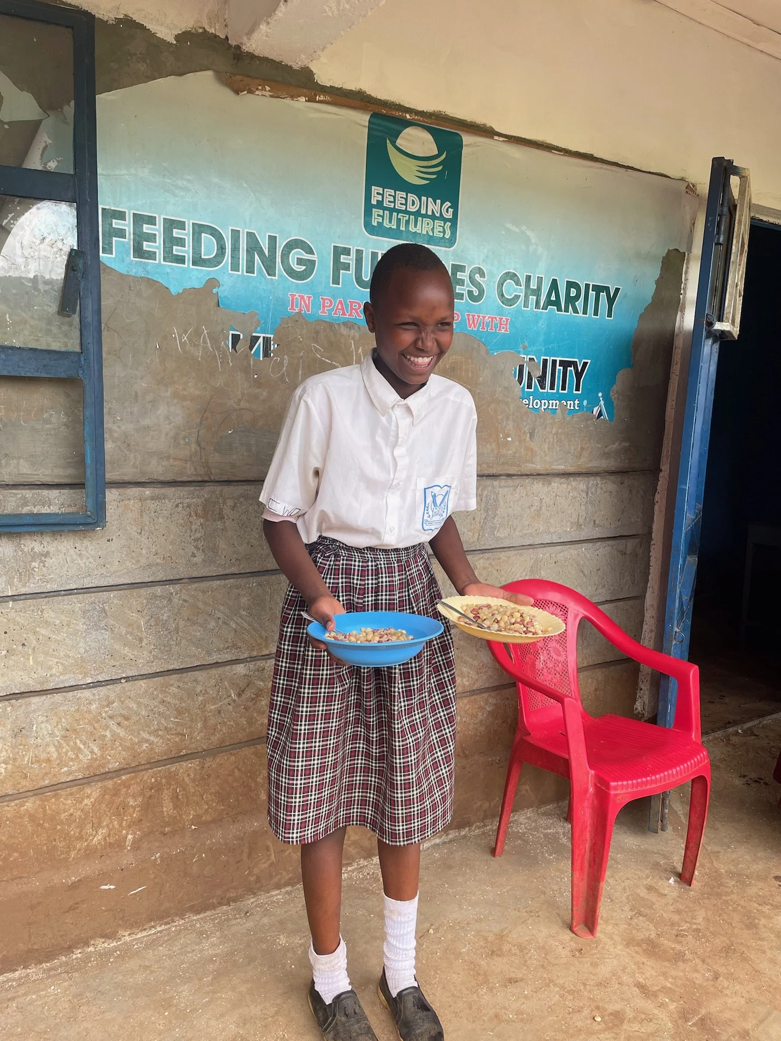 A young girl in school uniform, smiling and holding two bowls of food, standing in front of a wall with a charity banner that reads 'Feeding Futures'.