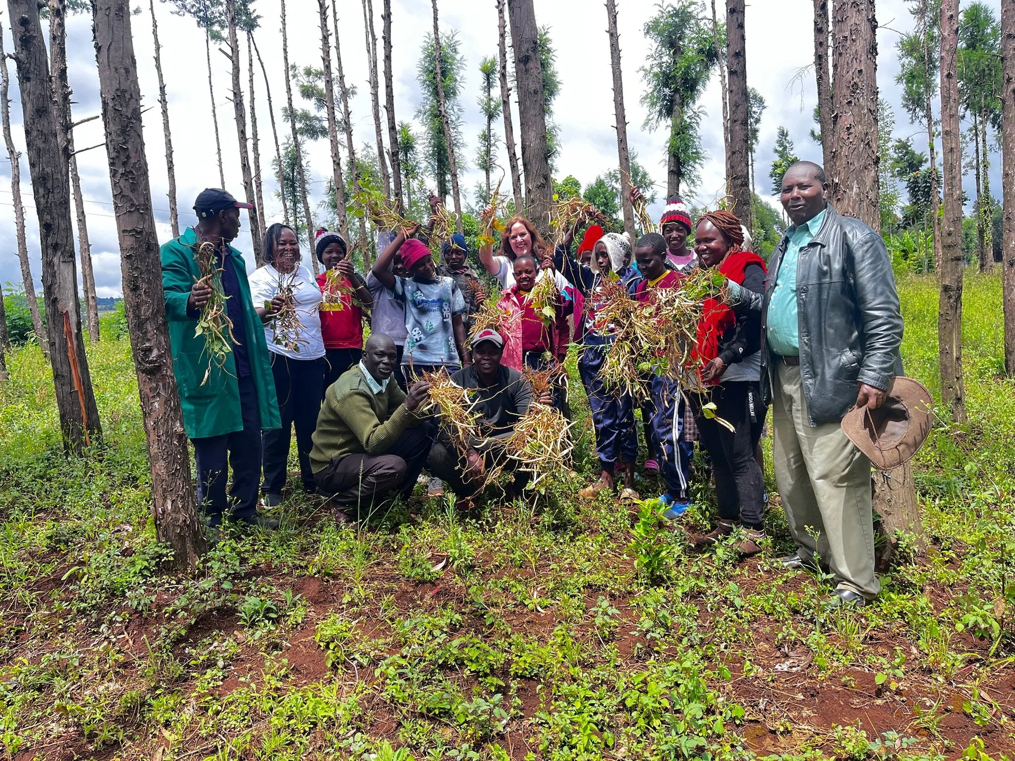 A group of people outdoors in a forest, holding plants and smiling at the camera. There are trees in the background and some are squatting or standing.