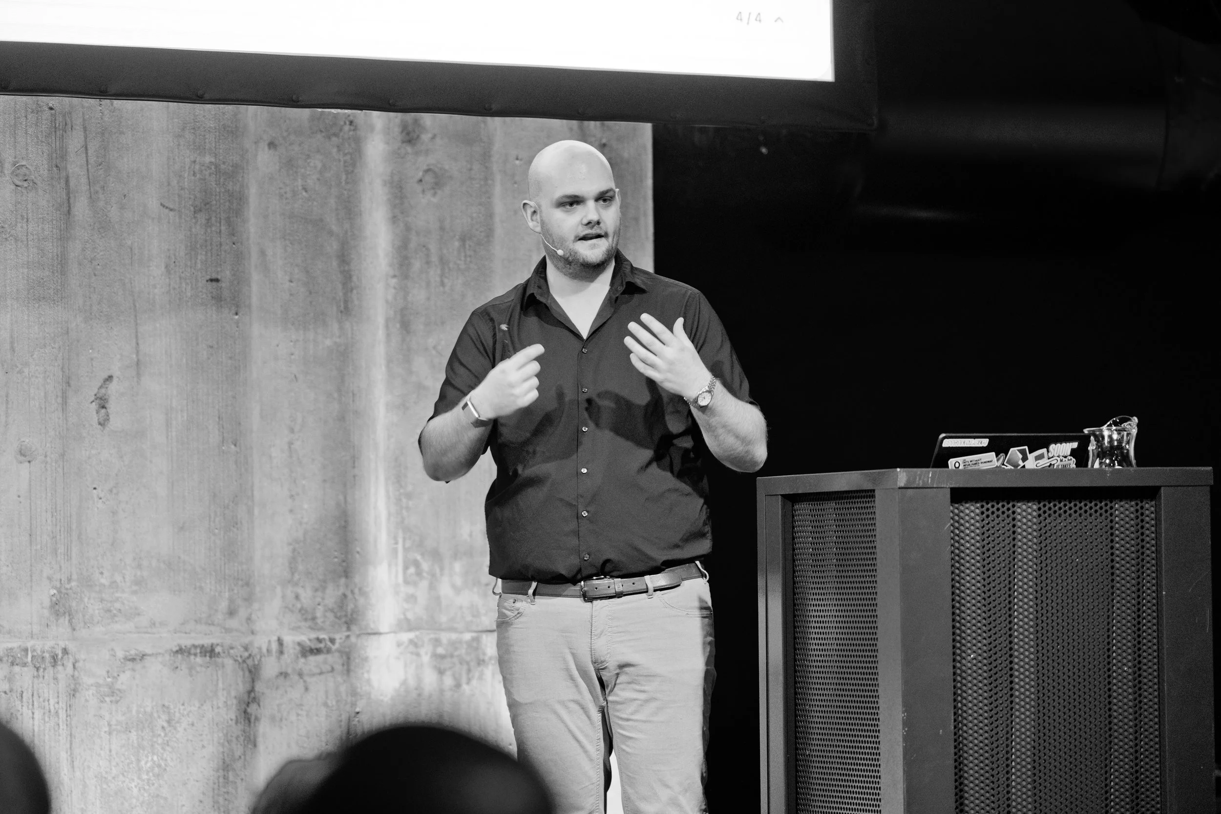 Ein Mann mit Glatze spricht vor einem Publikum. Er trägt ein schwarzes Hemd, eine Armbanduhr und einen Ring, und steht neben einem Pult, auf dem ein Laptop mit mehreren Stickern steht. Die Szene ist in Schwarz-Weiß. Hinter ihm eine graue Betonwand.