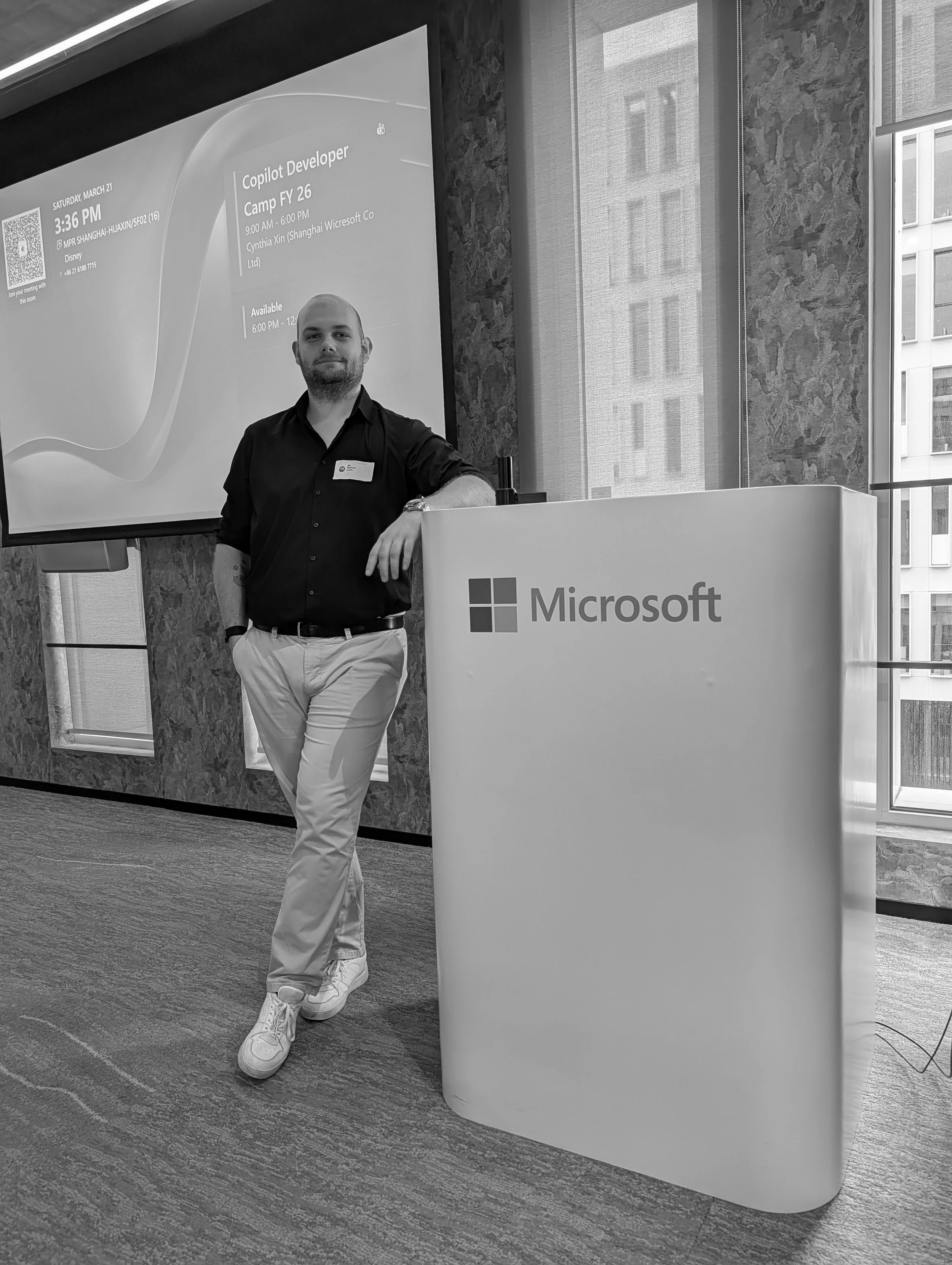 Mann steht vor einem Microsoft-Stand bei einer Veranstaltung, mit einem großen Bildschirm im Hintergrund, auf dem Angaben zu einem Programm und einer Person zu sehen sind.