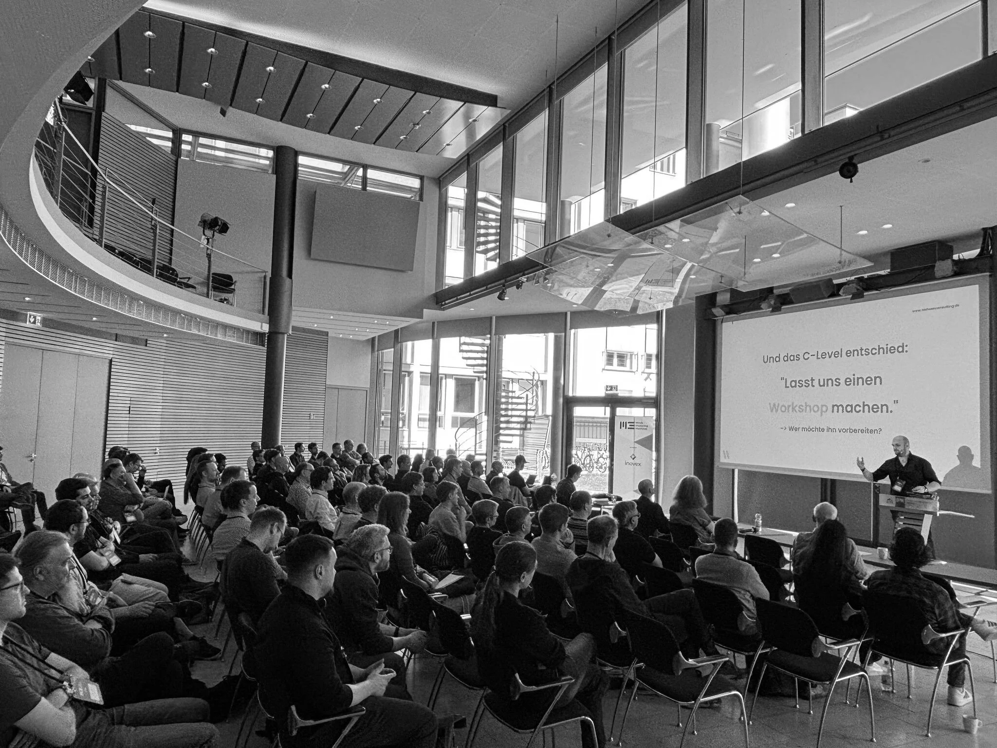 Ein großer Konferenzraum mit vielen Menschen, die einem Vortrag zuhören. Es gibt große Fenster, die viel Tageslicht hereinlassen, und einen Bildschirm an der Wand.