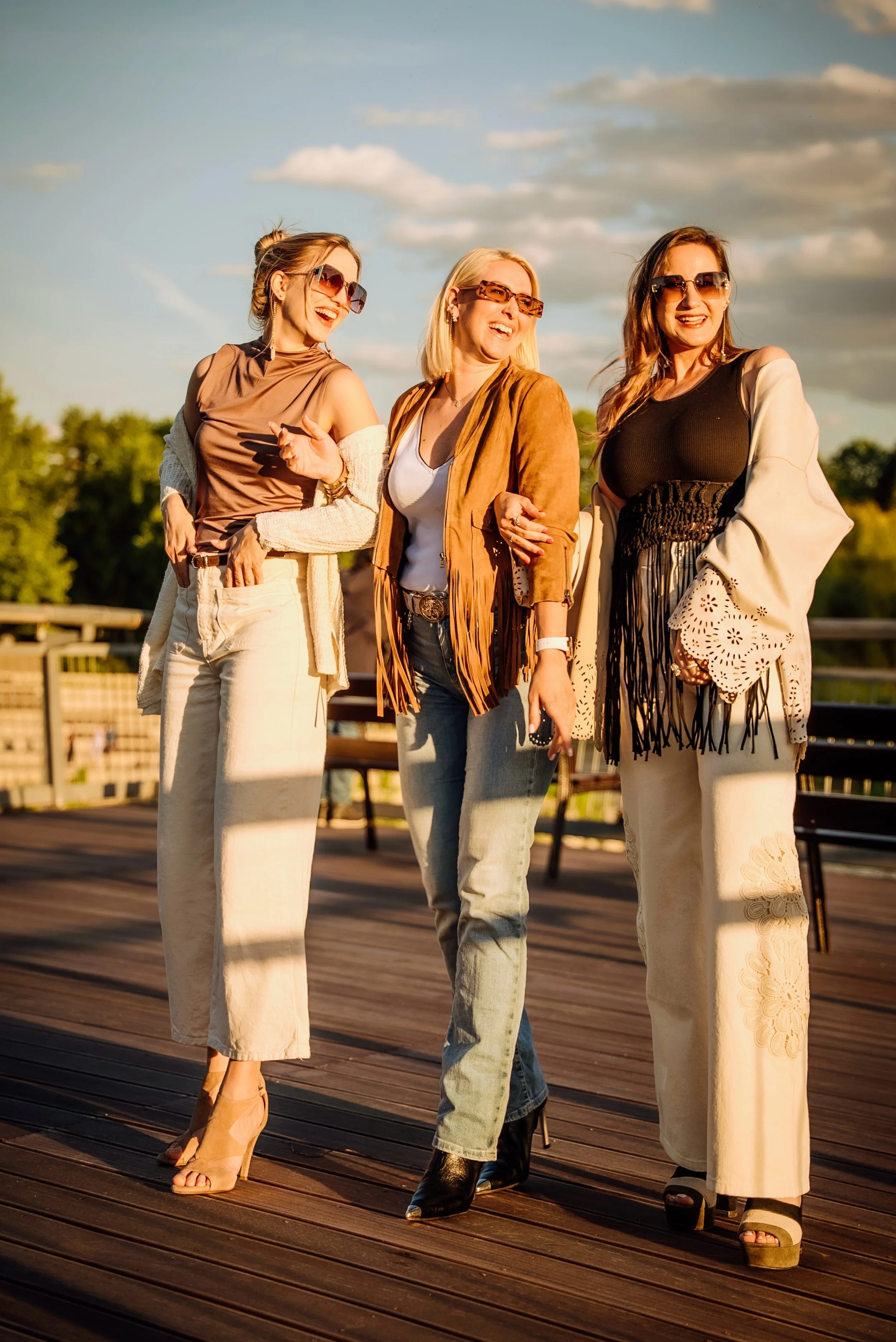 Three women walking outdoors during sunset, laughing and wearing fashionable, stylish outfits and sunglasses.