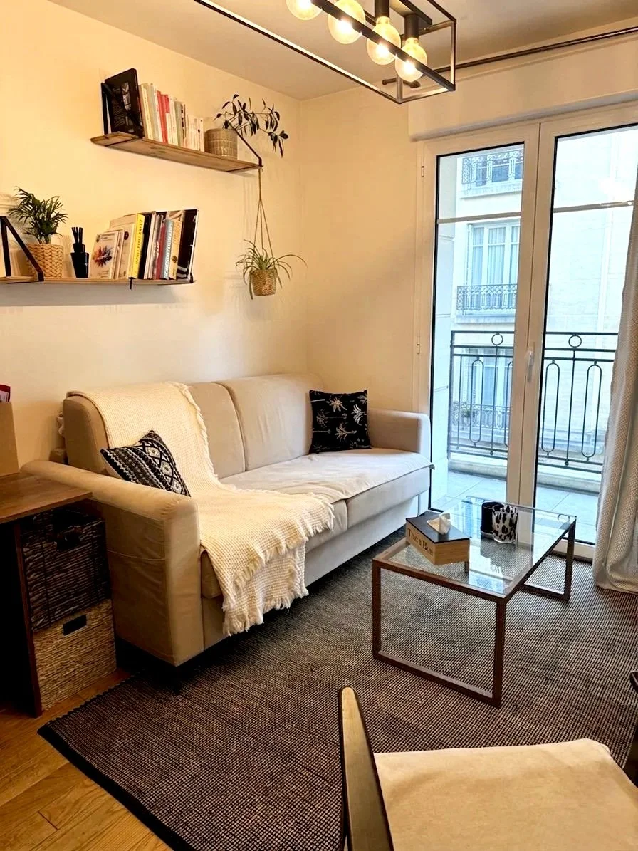 Salon cosy avec un canapé beige, deux coussins noirs, une table basse en verre, une étagère avec livres et plantes, une grande fenêtre avec un balcon.