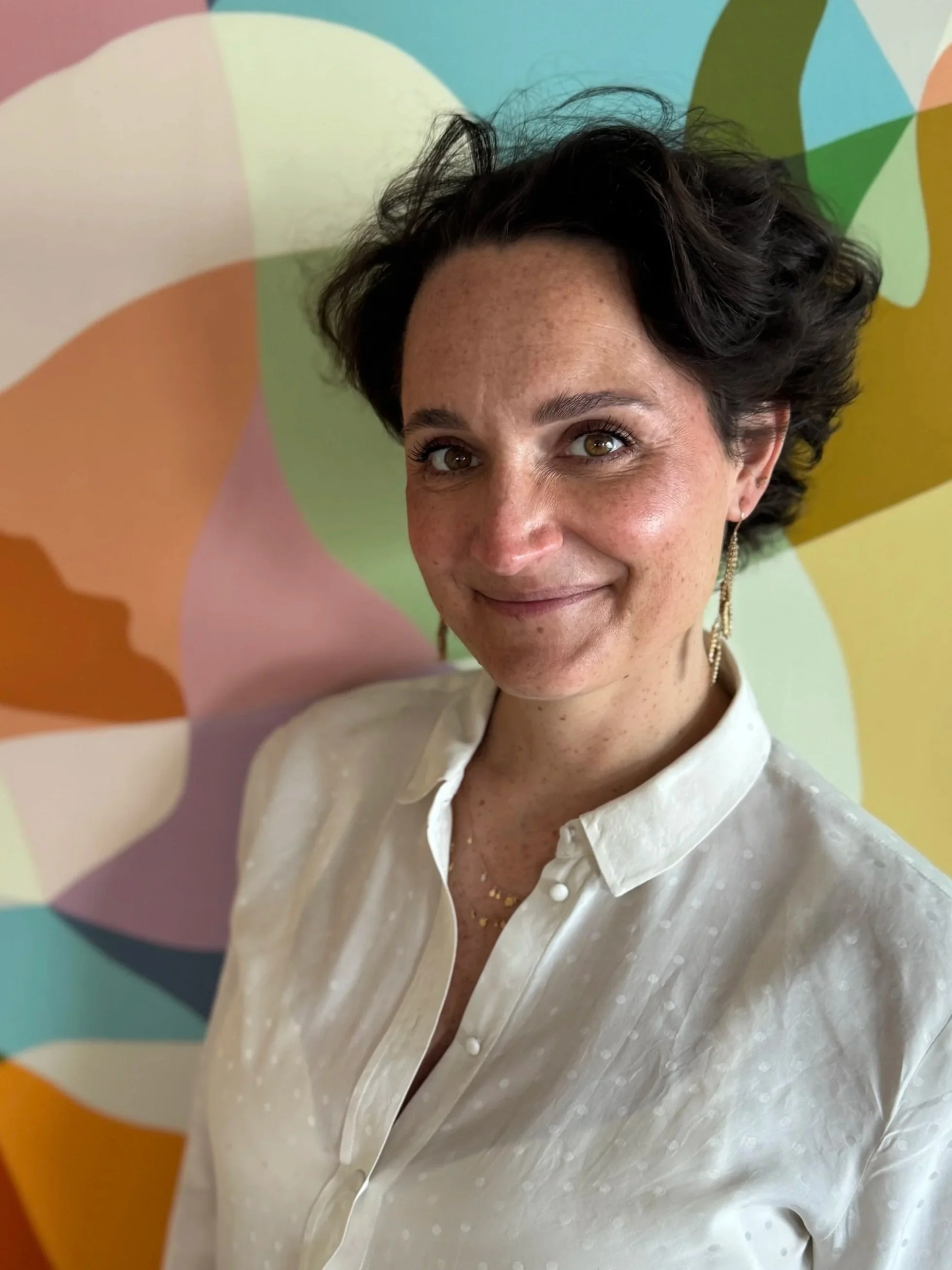 Portrait d'une femme souriante avec des cheveux courts et bouclés, portant une chemise blanche et des boucles d'oreilles longues, contre un fond coloré abstrait.