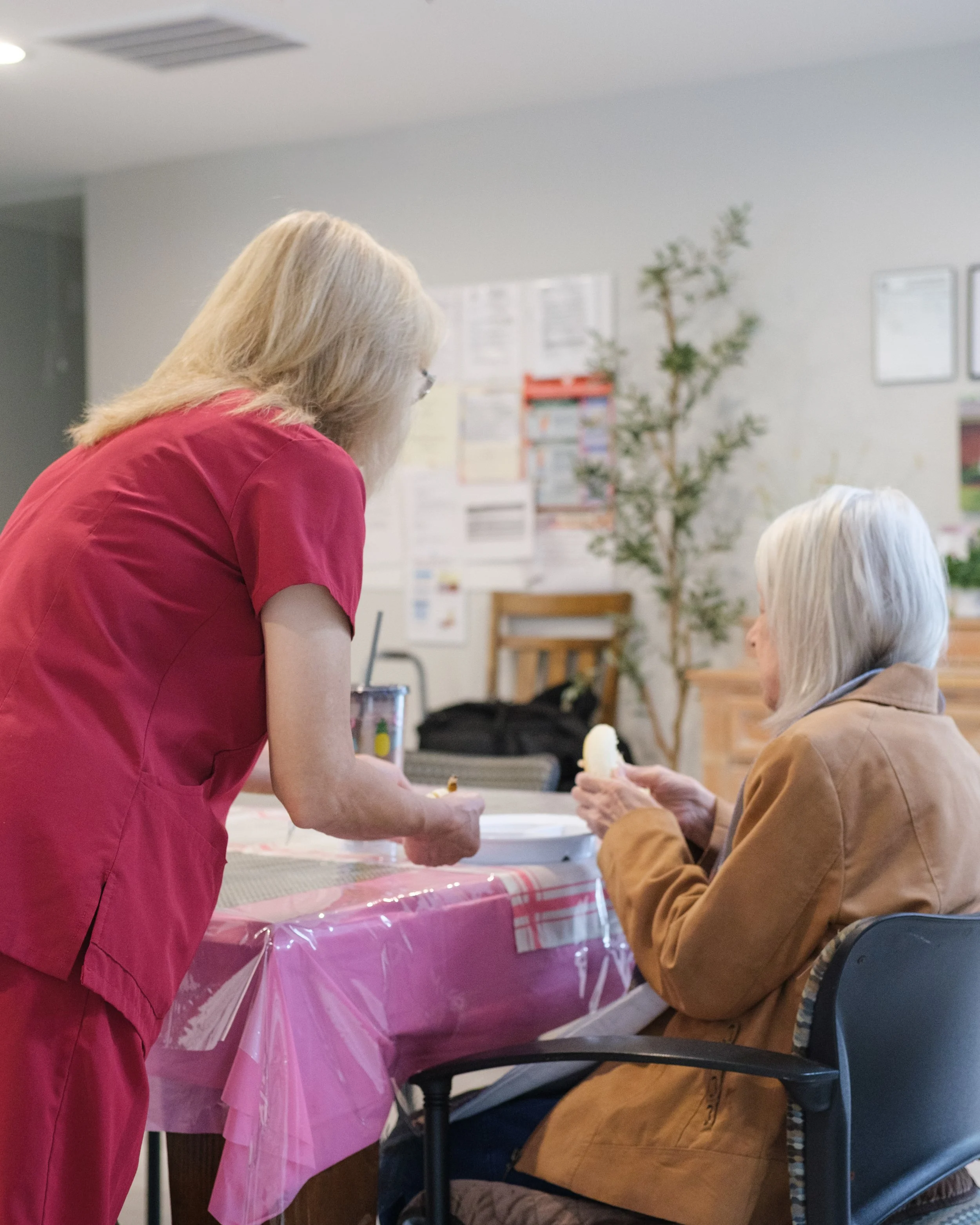 A woman in pink scrubs is standing and talking to an elderly woman with long white hair, who is sitting in a chair. The elderly woman is holding a snow cone. There is a table covered with a pink plastic tablecloth between them, and in the background,