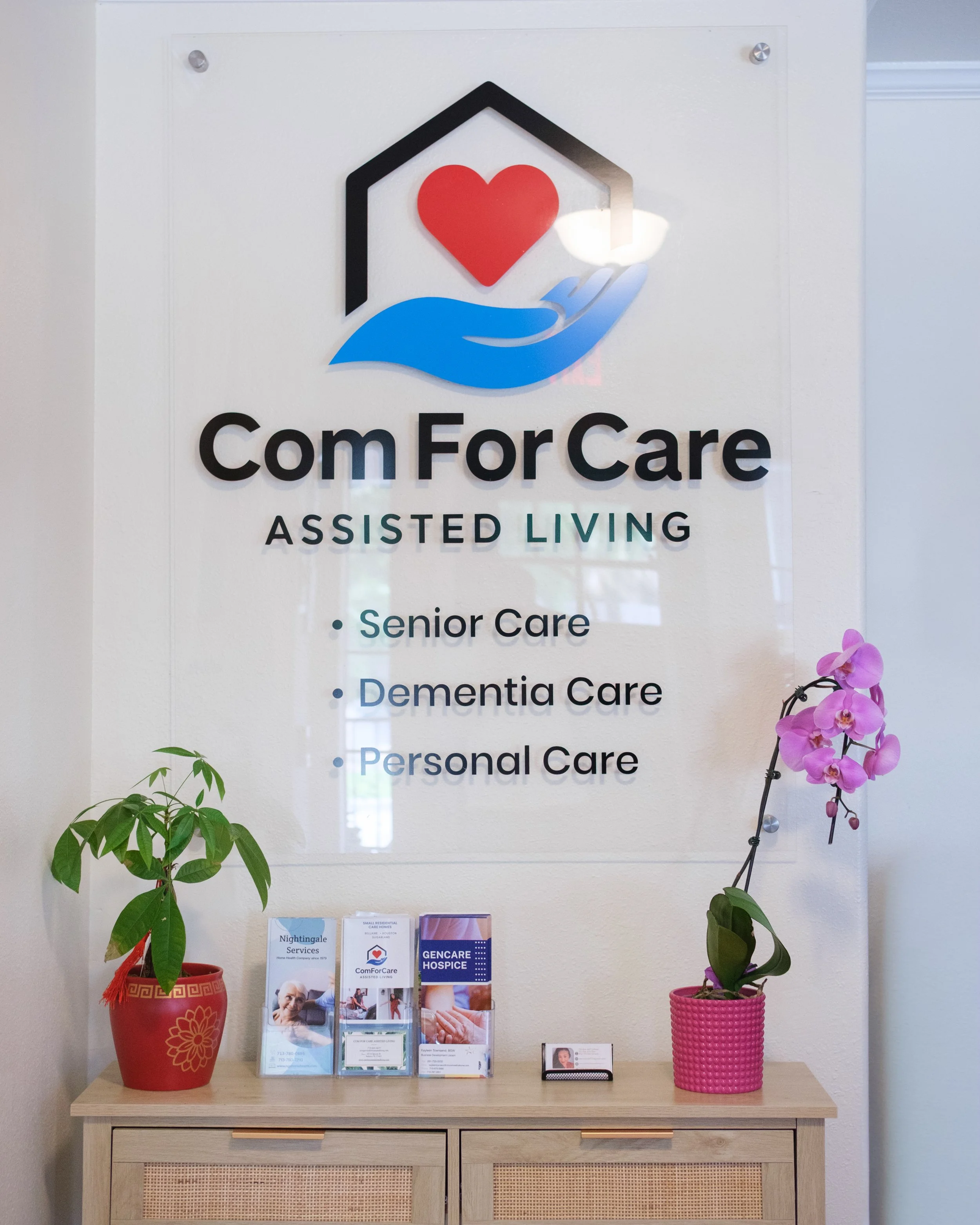 A wooden table with two potted plants, a red one on the left with green leaves and a decorative gold pattern, and a pink one on the right with purple orchids. Above the table, there is a white sign with the logo and name of a senior care organization