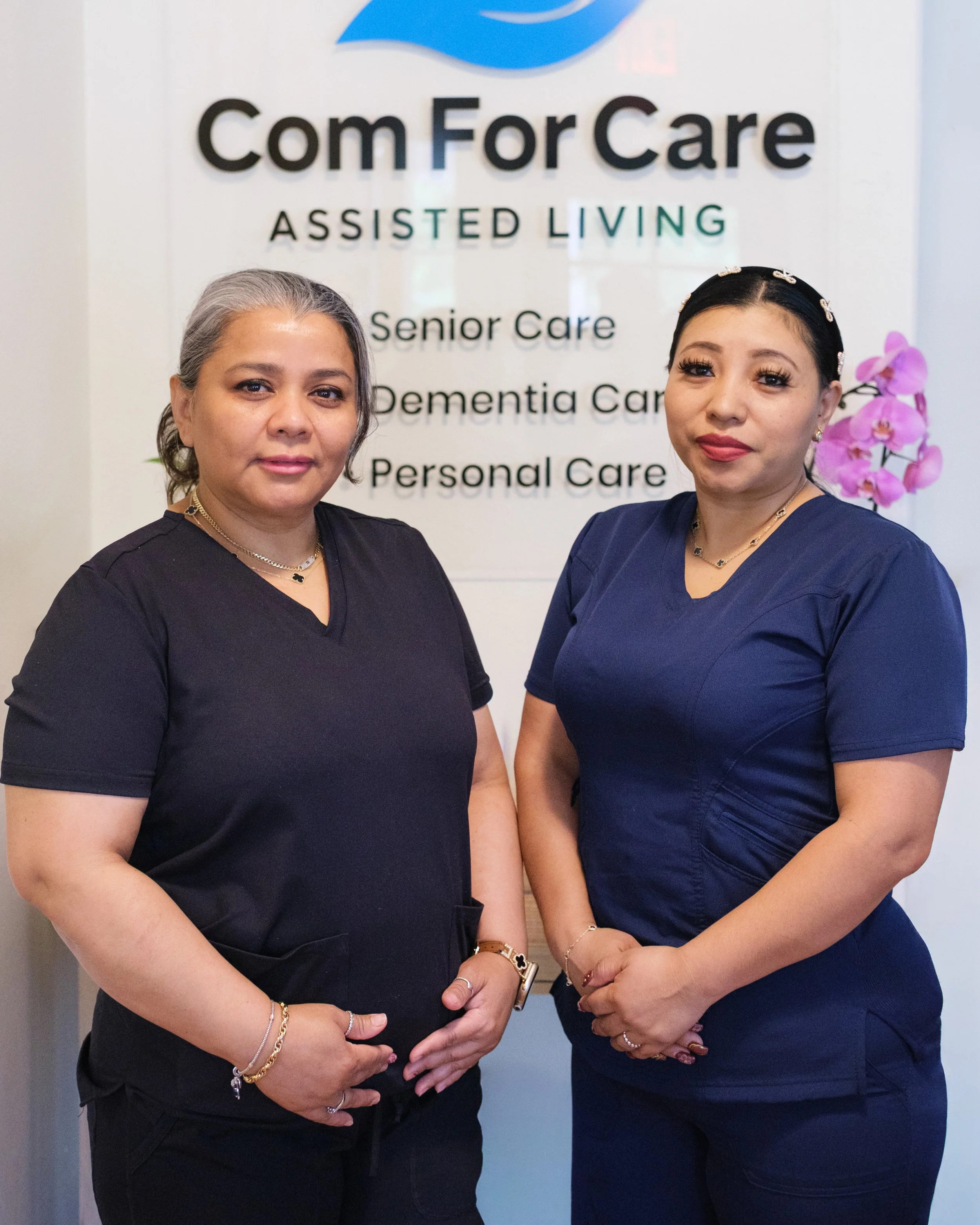 Two women in medical scrubs standing in front of a sign that reads "ComForCare Assisted Living, Senior Care, Dementia Care, Personal Care," with a pink orchid plant nearby.