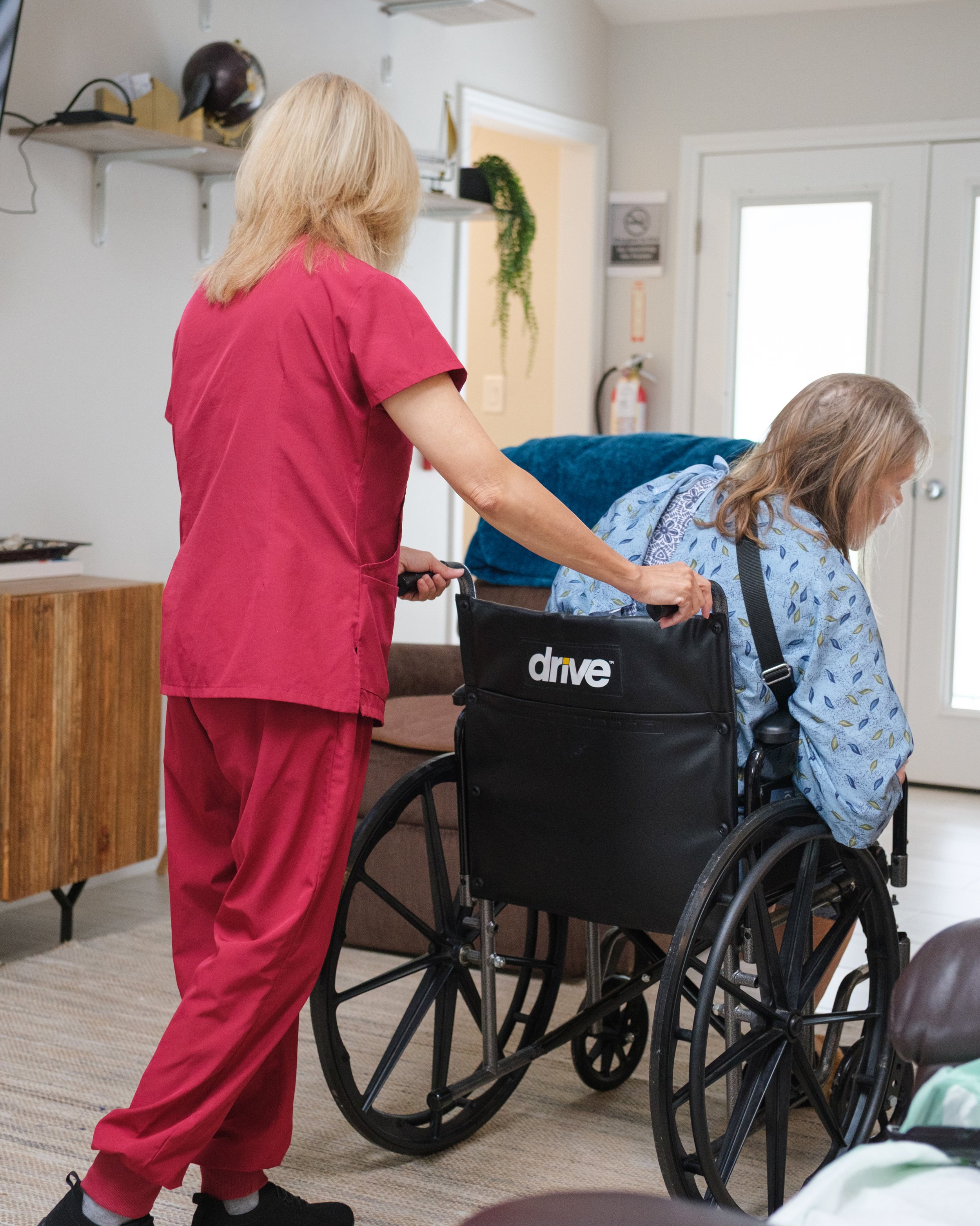 A healthcare worker in pink scrubs assisting a woman in a wheelchair with light brown hair in a medical setting.