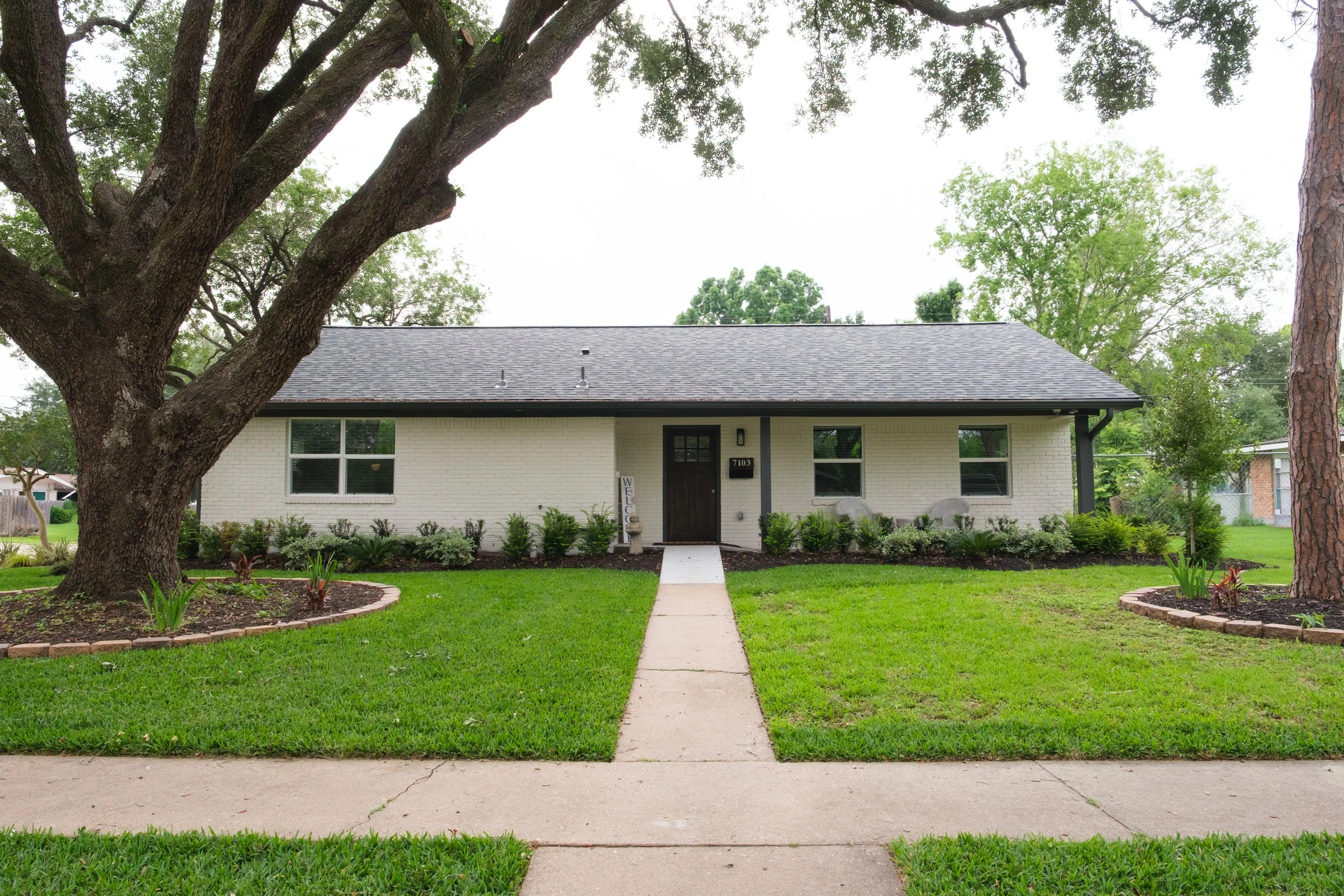 Front view of a white house with black door, surrounded by a green lawn and garden beds, with large trees in the yard and neighboring houses in the background.
