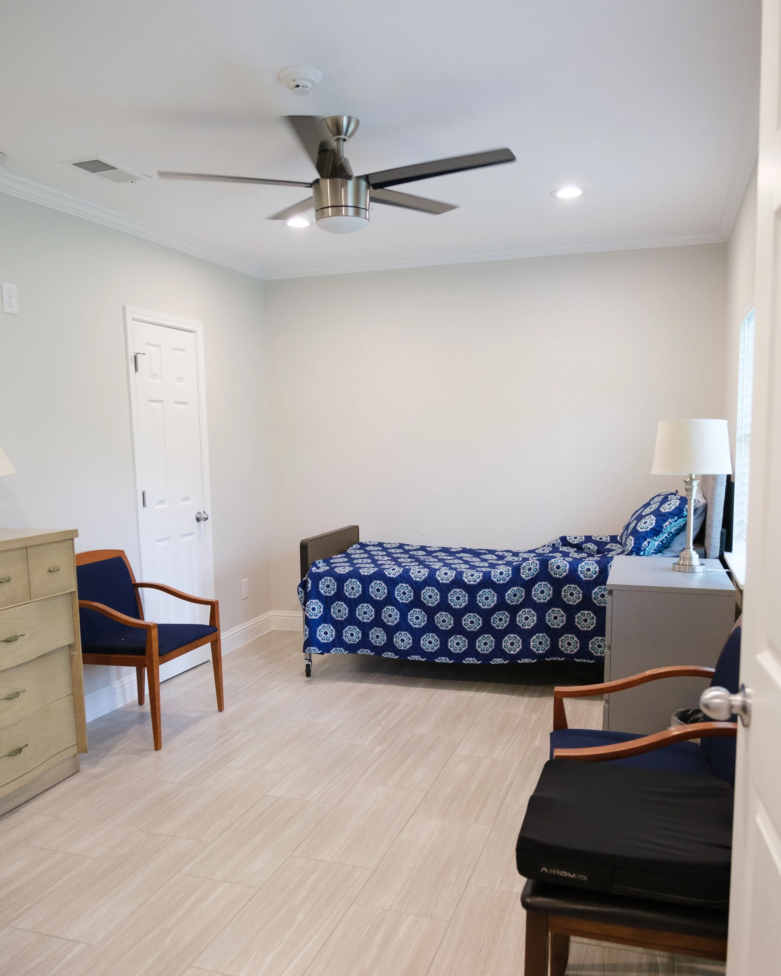 A simple bedroom with a ceiling fan, two ceiling lights, a white door, a beige dresser, chairs, a bed with a blue patterned bedspread, a nightstand with a lamp, and a window with blinds.