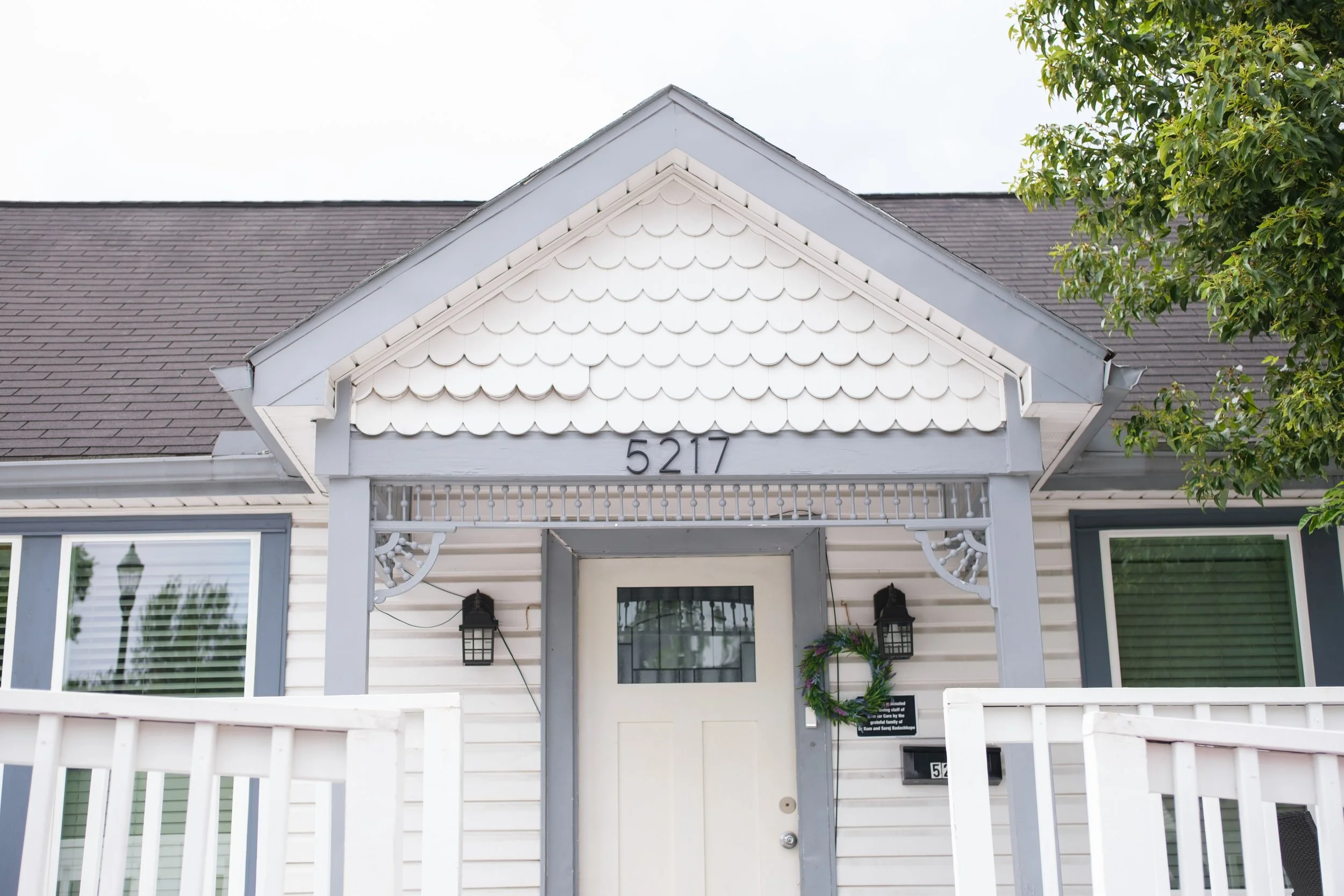 Front view of a house with white siding, a covered porch, and a gable roof with gray shingles. The house number 5217 is displayed above the porch. There is a white picket fence in the foreground, and a green tree to the right.